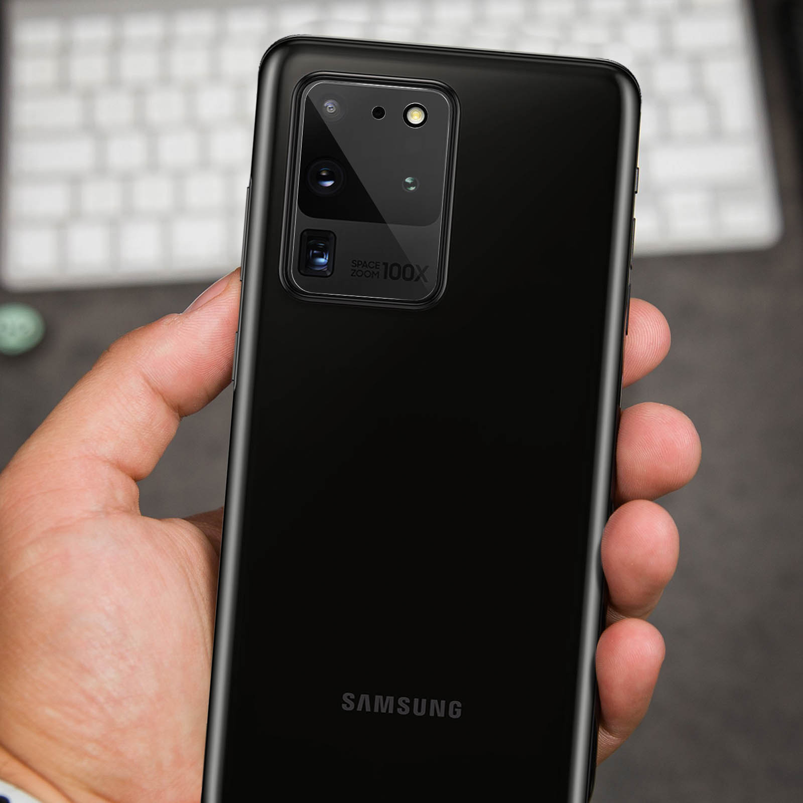 Mano che tiene un telefono Samsung nero. Il telefono ha più fotocamere e un'etichetta 'Space Zoom 100X'. Una tastiera sullo sfondo.