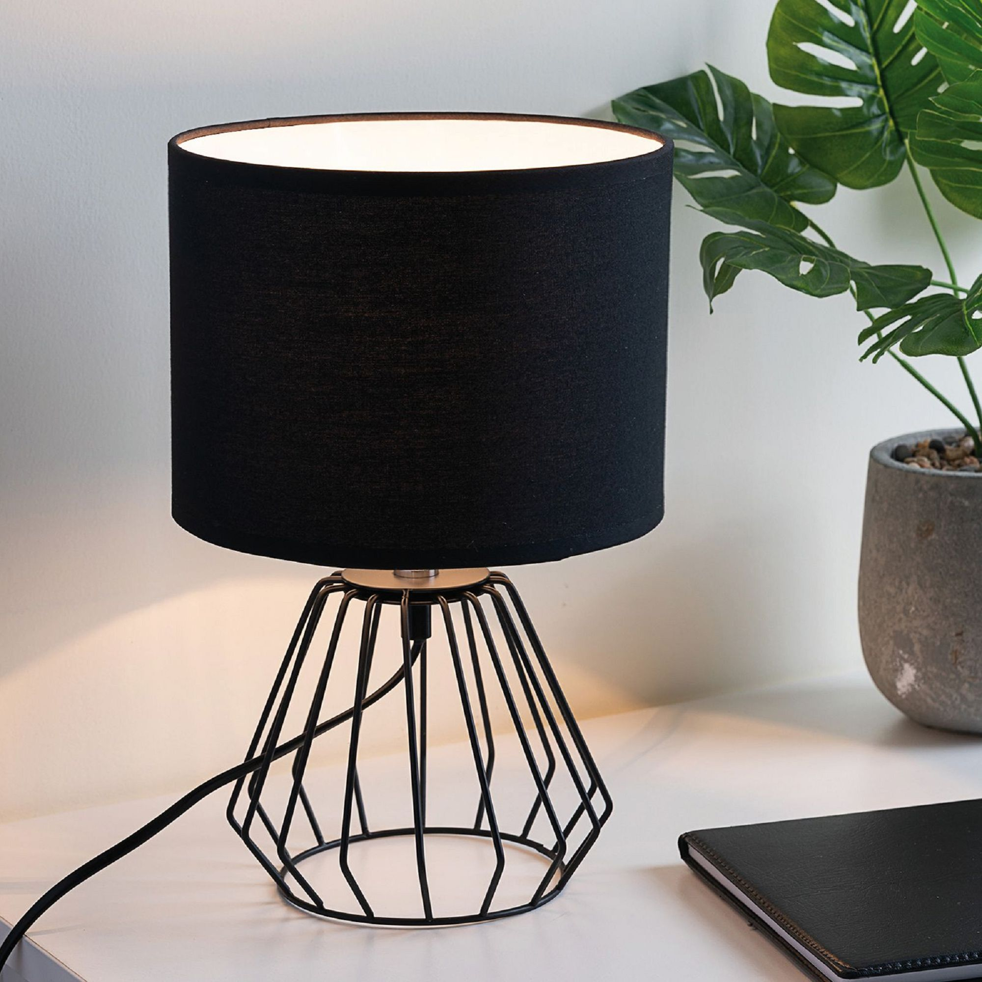 Czarna lampa z abażurem i geometryczną podstawą, na białym stole, z czarnym notatnikiem.