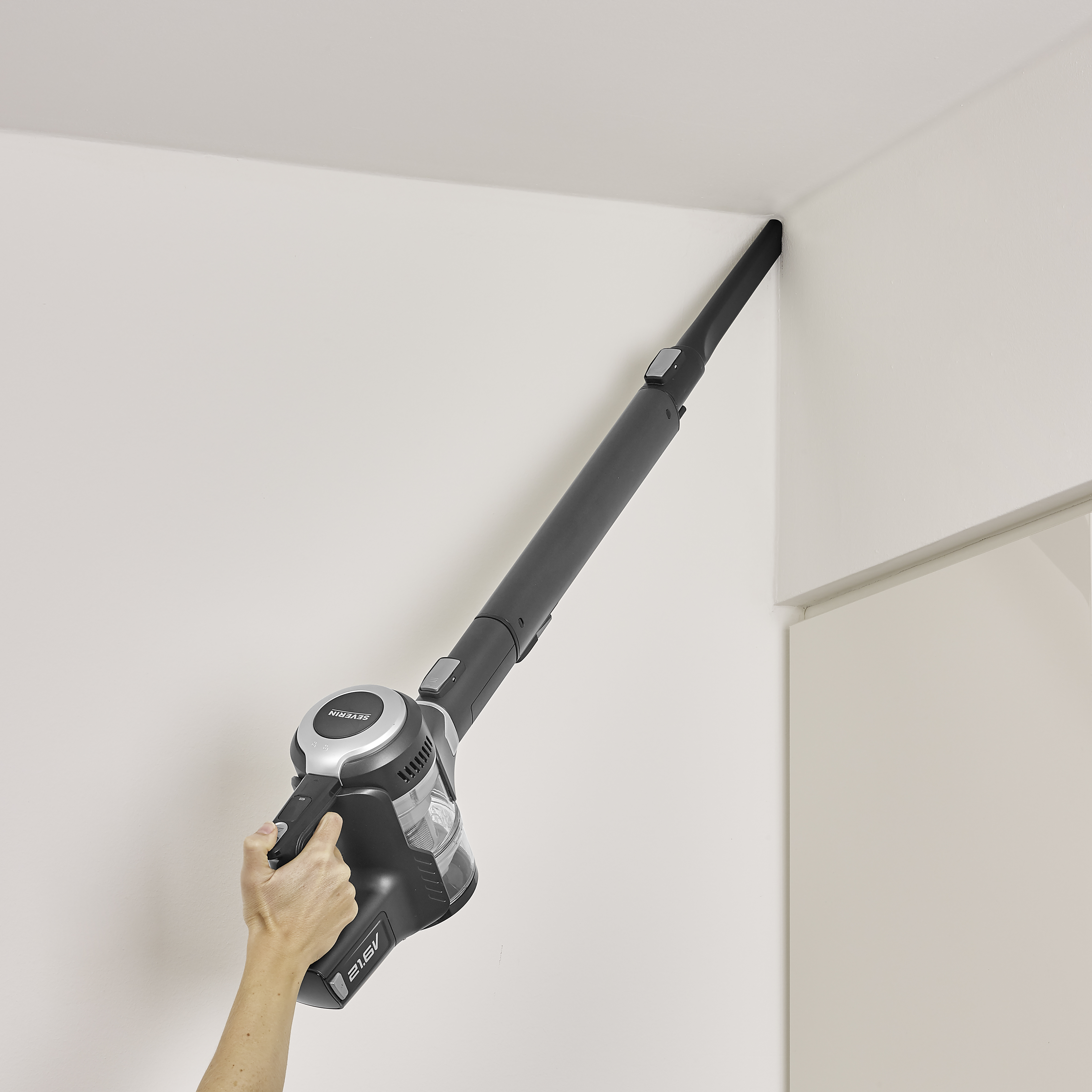 Una mano sujetando una aspiradora negra inalámbrica, limpiando una esquina de pared blanca.