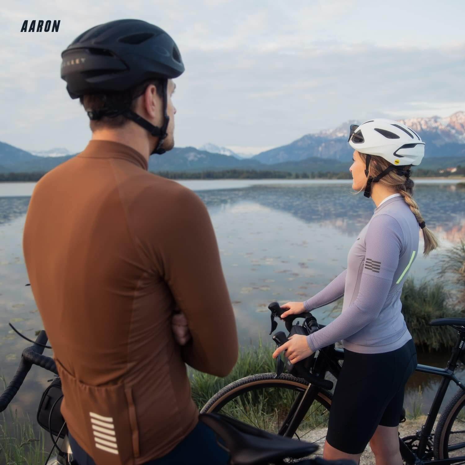 Zwei Radfahrer betrachten einen See und tragen Helme. Einer ist braun, der andere grau und weiß.