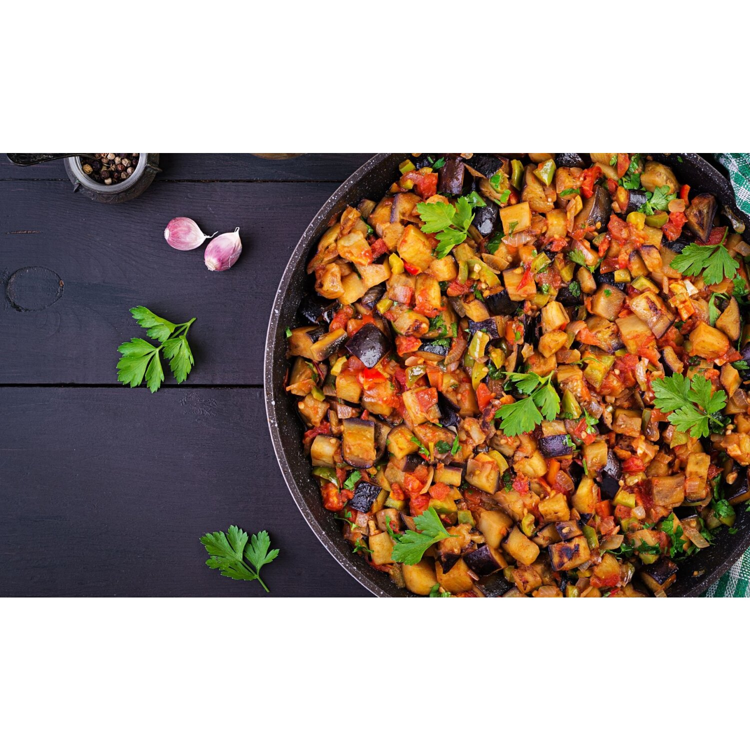 Pfanne mit gekochten, gewürfelten Auberginen, Tomaten und Kräutern auf schwarzem Holztisch.