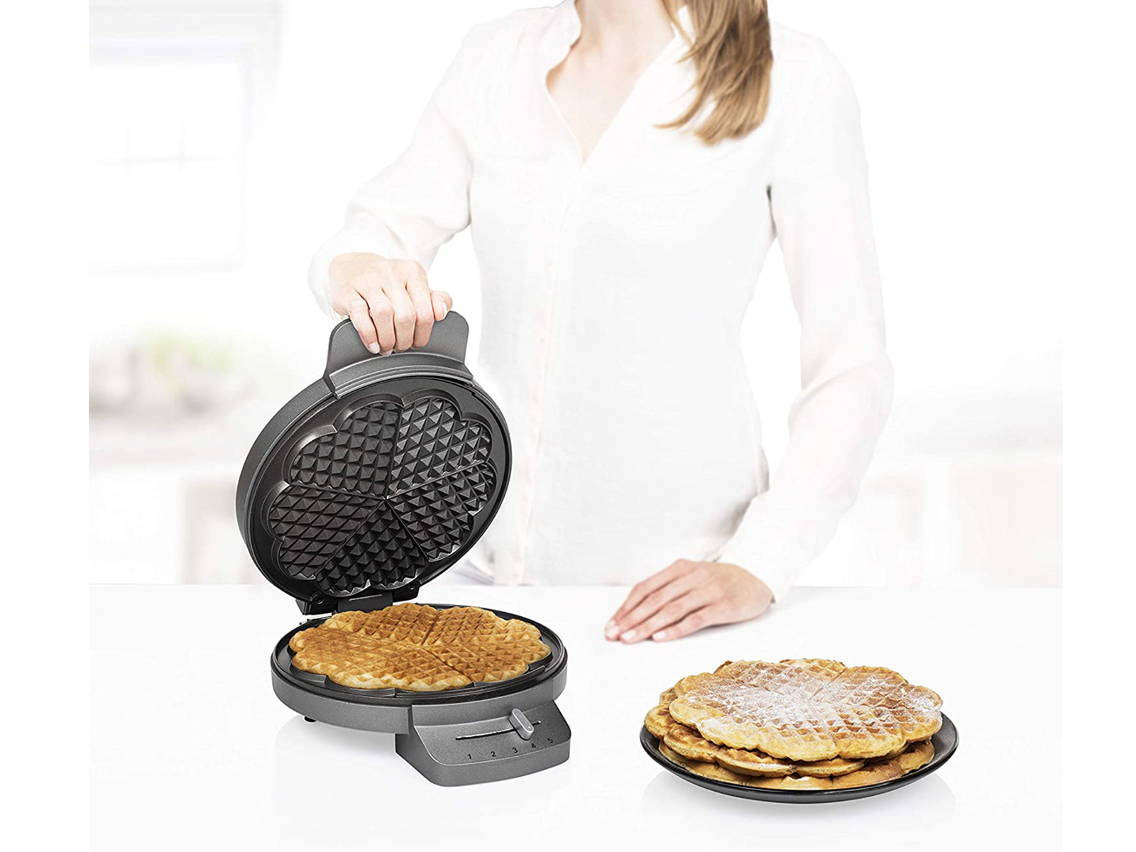 Eine Frau macht Waffeln in einem Waffeleisen. Waffeln sind auf einem Teller. Weitere Waffeln sind fertig.