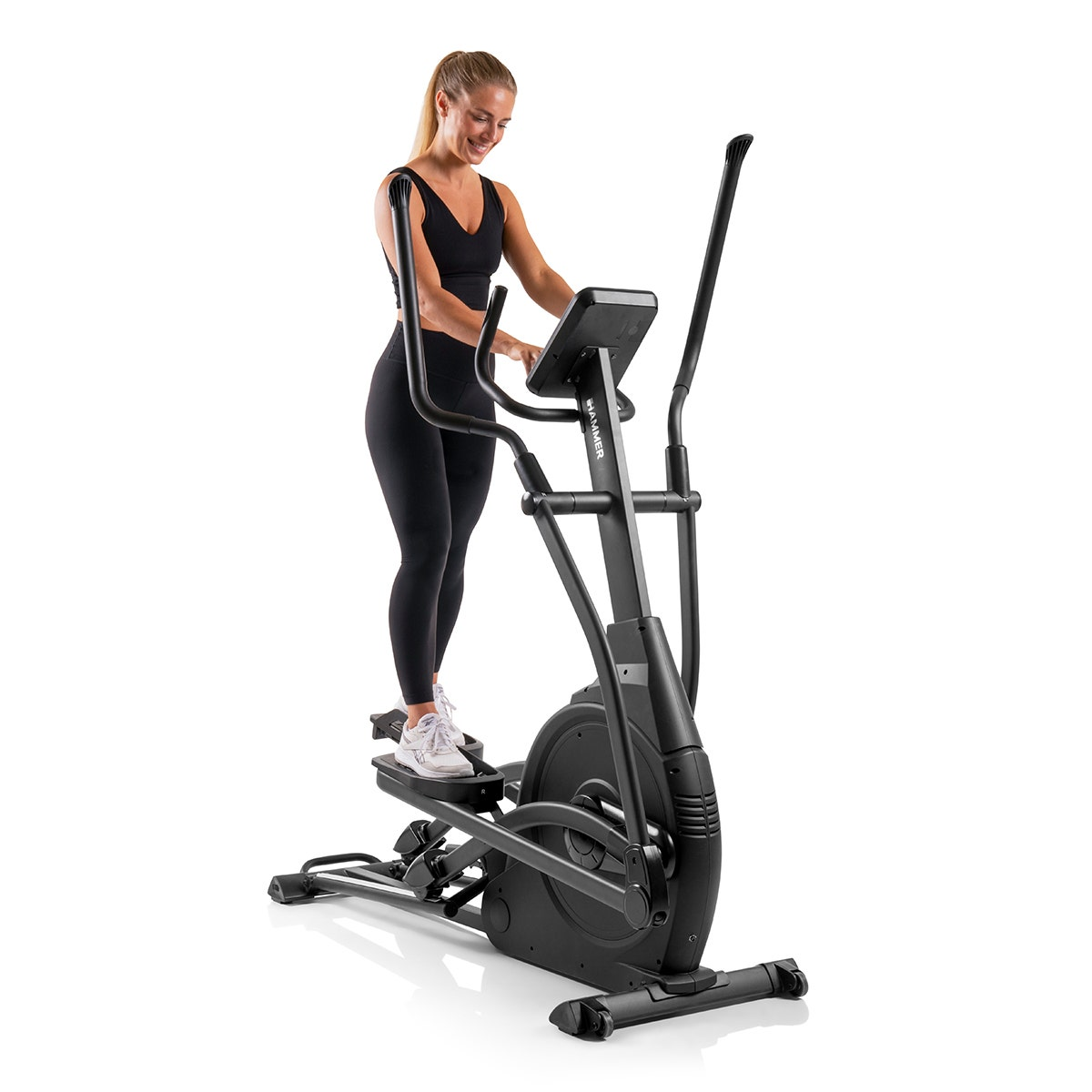 Eine Frau auf einem Crosstrainer. Sie lächelt, trägt schwarze Trainingskleidung, vor weißem Hintergrund.