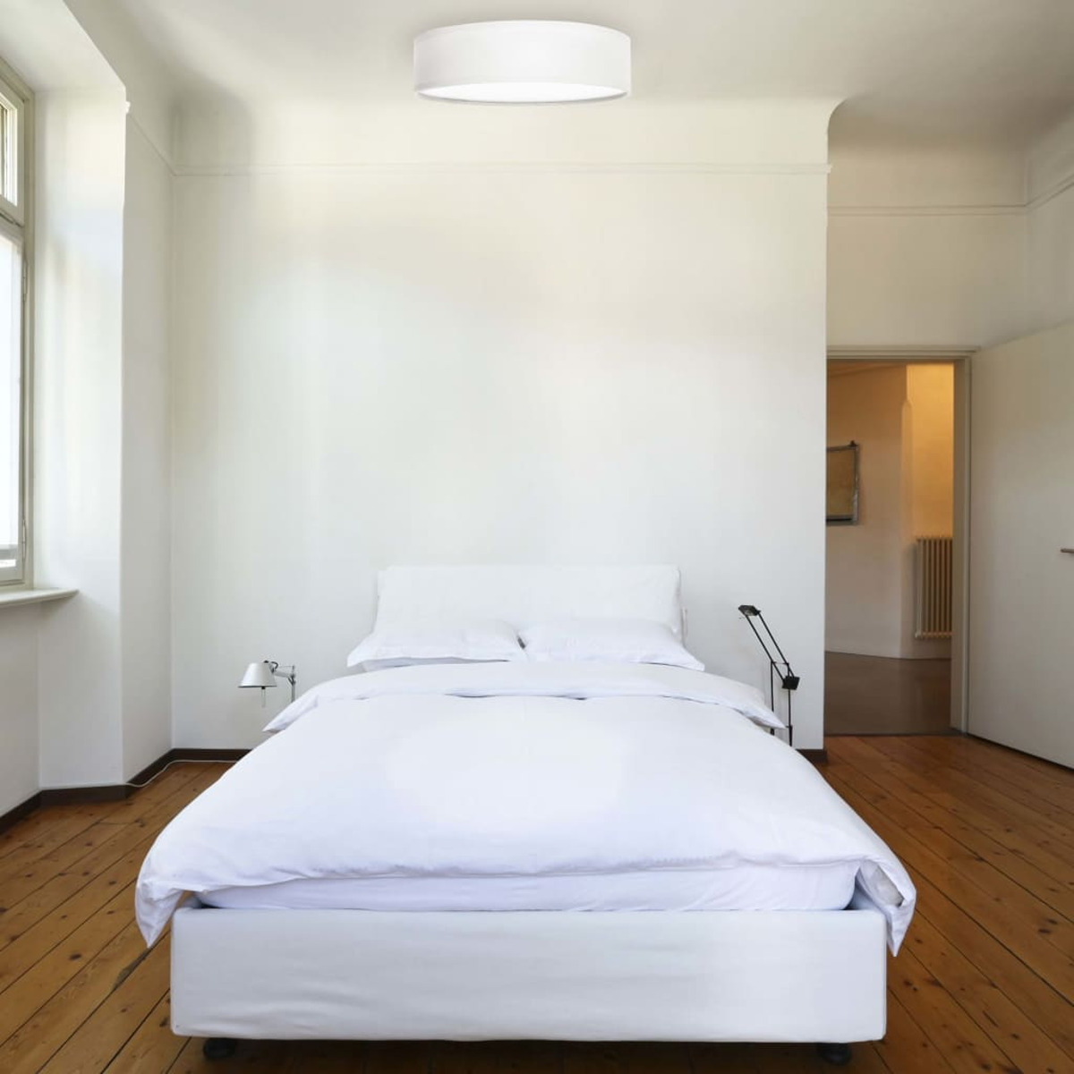 Una camera da letto bianca con letto, biancheria da letto bianca, comodino e luce a soffitto.