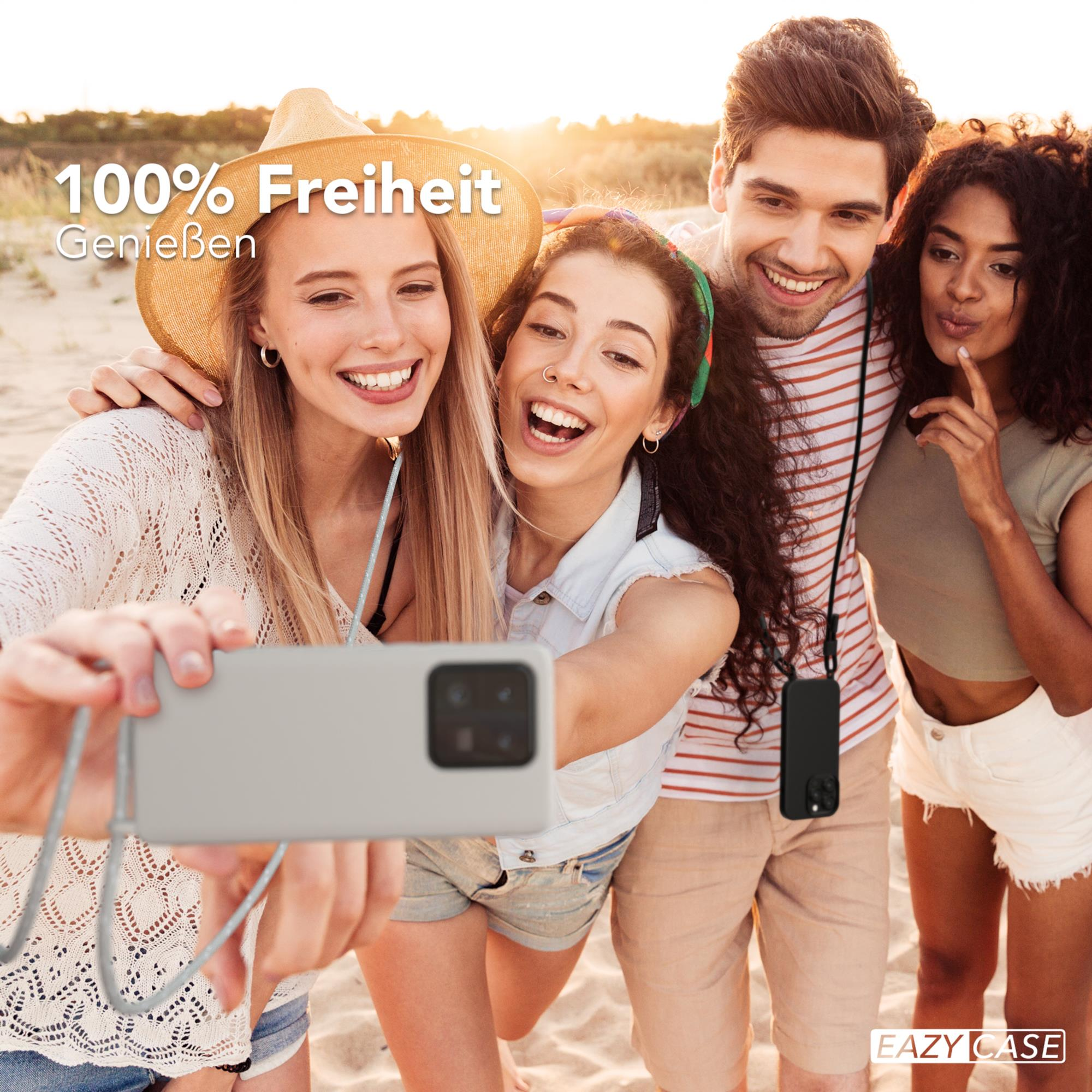 Vier Freunde am Strand lächeln, einer macht ein Selfie mit einem grauen Telefon. Sand und Himmel im Hintergrund.