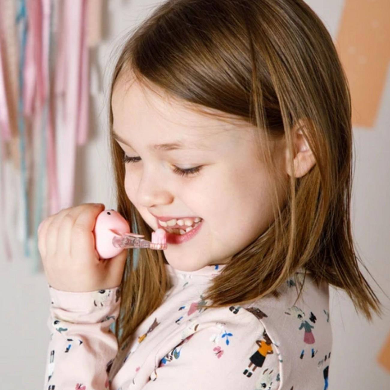 Una bambina si lava i denti con uno spazzolino rosa, sorridendo, sfondo rosa chiaro.