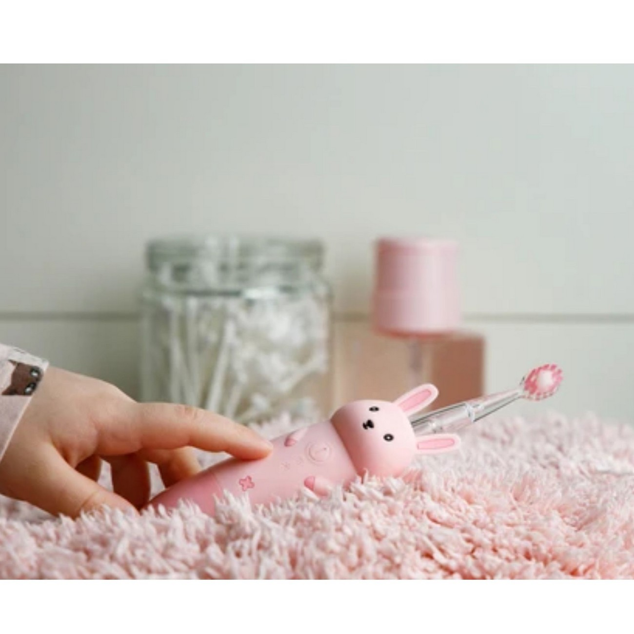 Una mano di bambino tiene uno spazzolino elettrico rosa a forma di coniglio su un tappeto rosa.