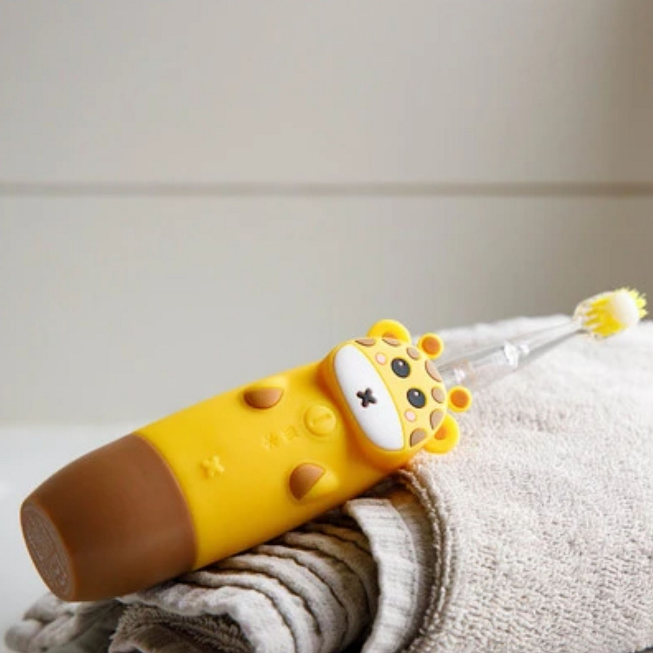 Cepillo de dientes amarillo con diseño de jirafa. Está sobre una superficie blanca con una toalla.