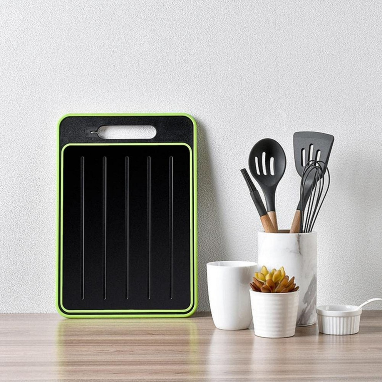 Tabla de cortar negra con borde verde, junto a utensilios y una pequeña planta.
