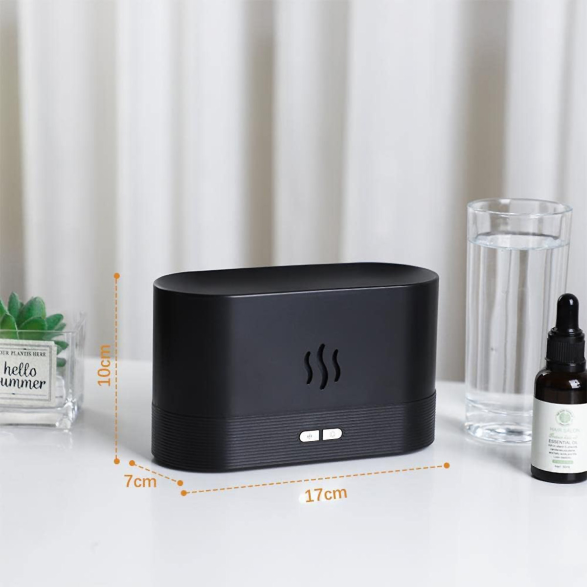 Difusor de aroma negro con dimensiones. Vaso de agua y botella de aceite sobre mesa blanca.