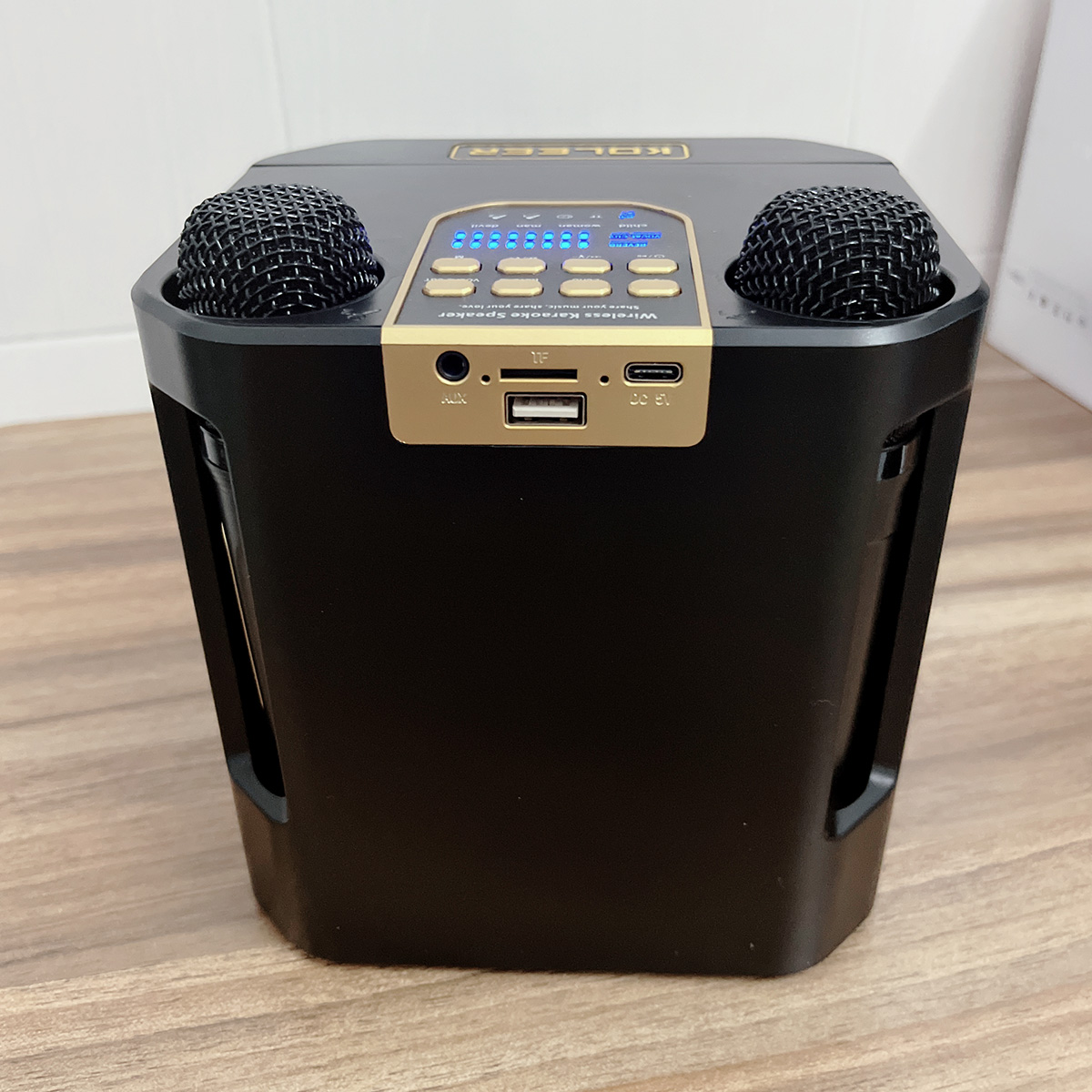 Máquina de karaoke negra con dos micrófonos, botones y puertos. Fondo de suelo de madera.