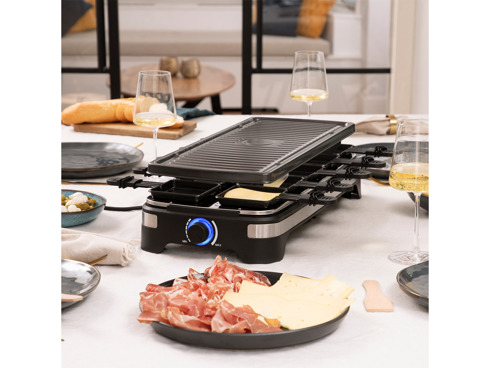 Tisch für Raclette mit Essen und Weingläsern, mit verschwommenem Hintergrund.