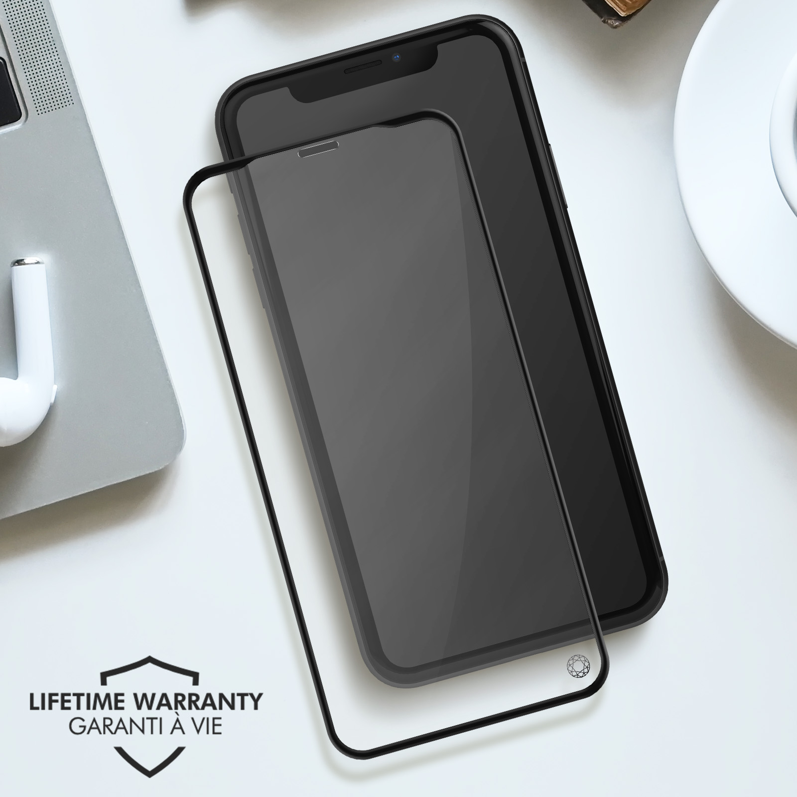 A phone and a screen protector are on a white surface. A laptop and a pair of earbuds are in the background.