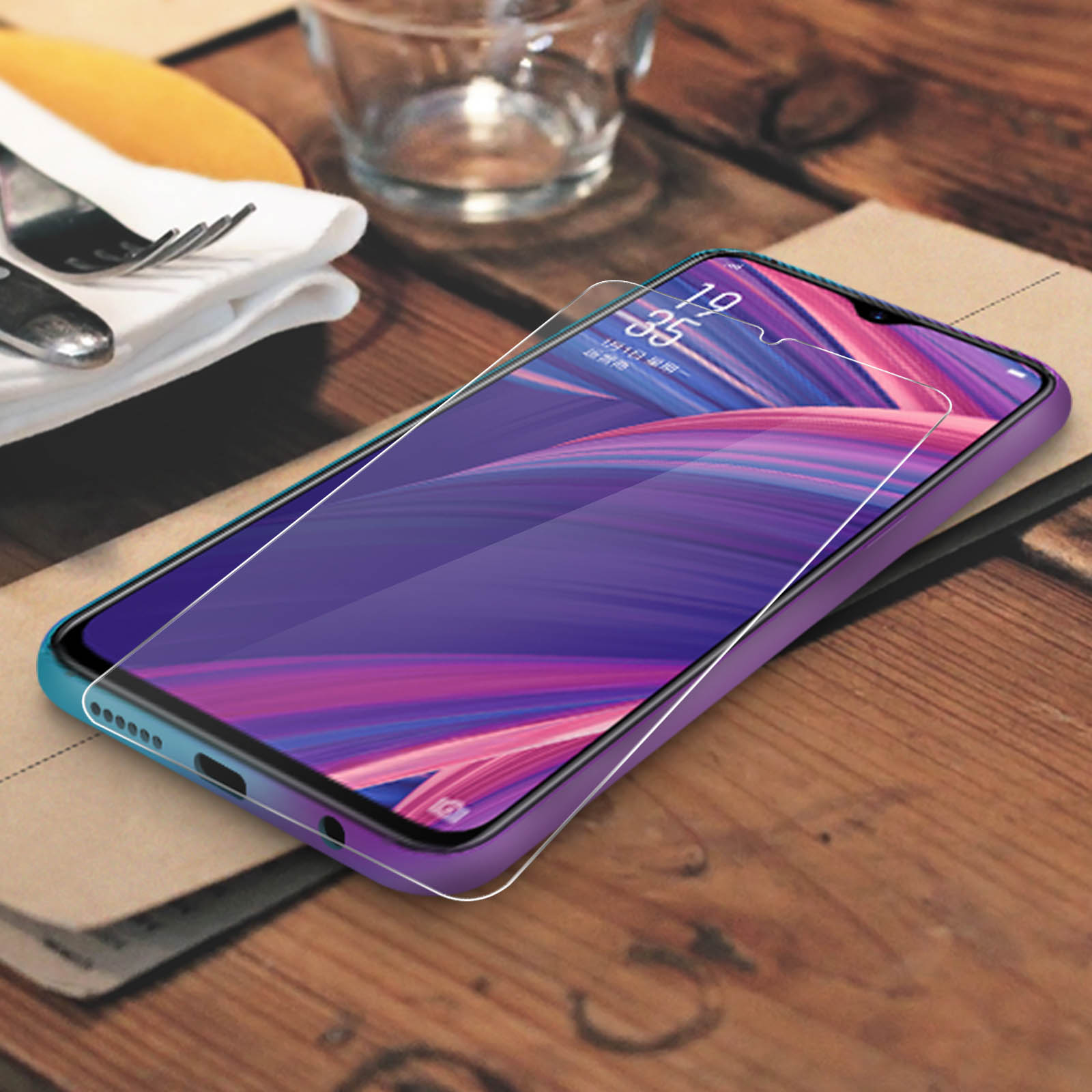 Uno smartphone con custodia viola è appoggiato su un tavolo di legno. Lo schermo mostra un design astratto colorato. Posate e bicchiere visibili.