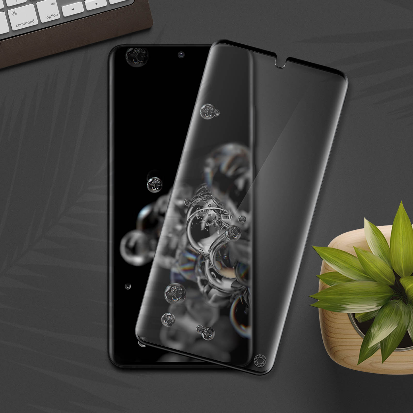 Due smartphone con protezioni schermo, una pianta e una tastiera su superficie scura. Bolle riflesse.