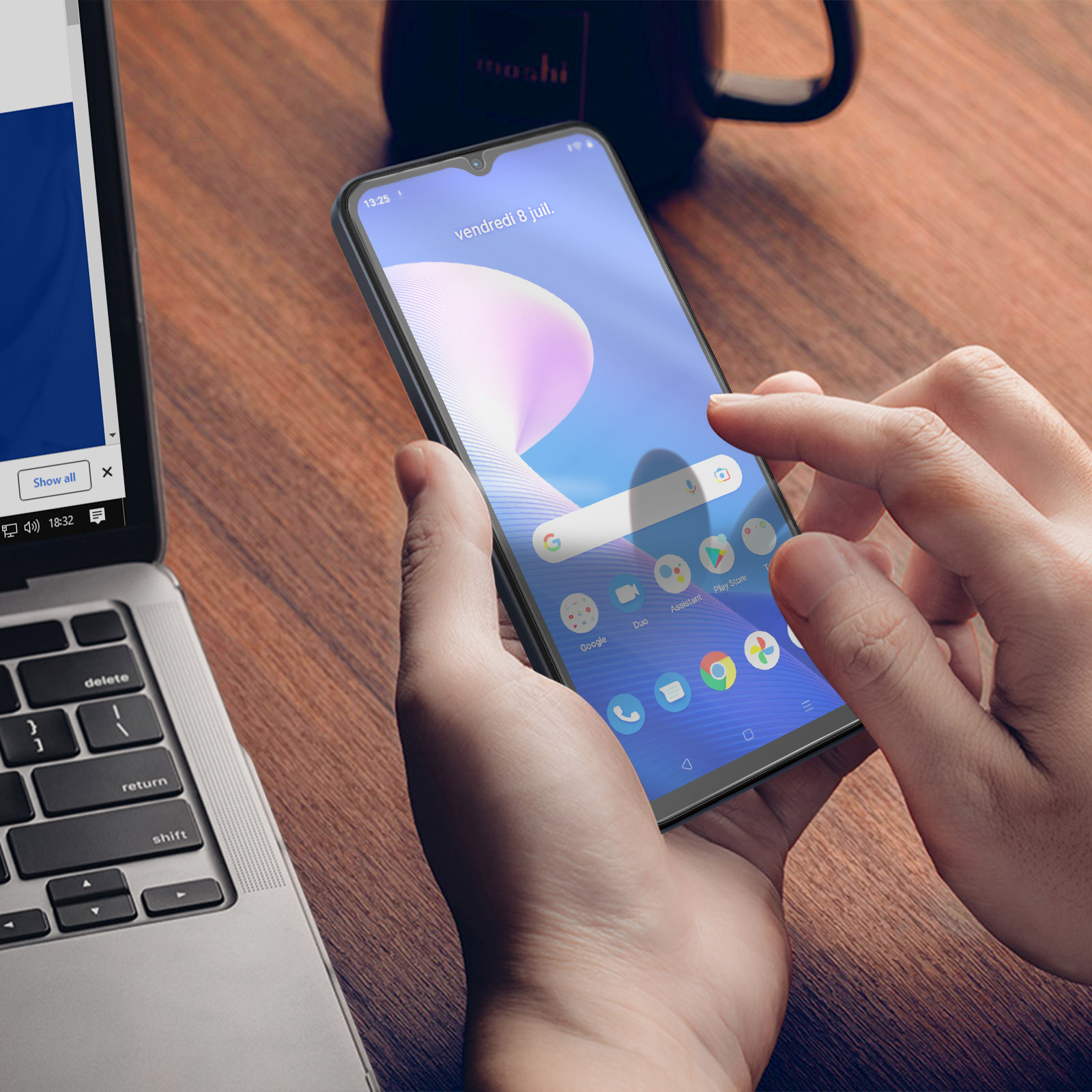 Une main touche l'écran d'un smartphone affichant des icônes d'applications. Un ordinateur portable et une tasse en arrière-plan.