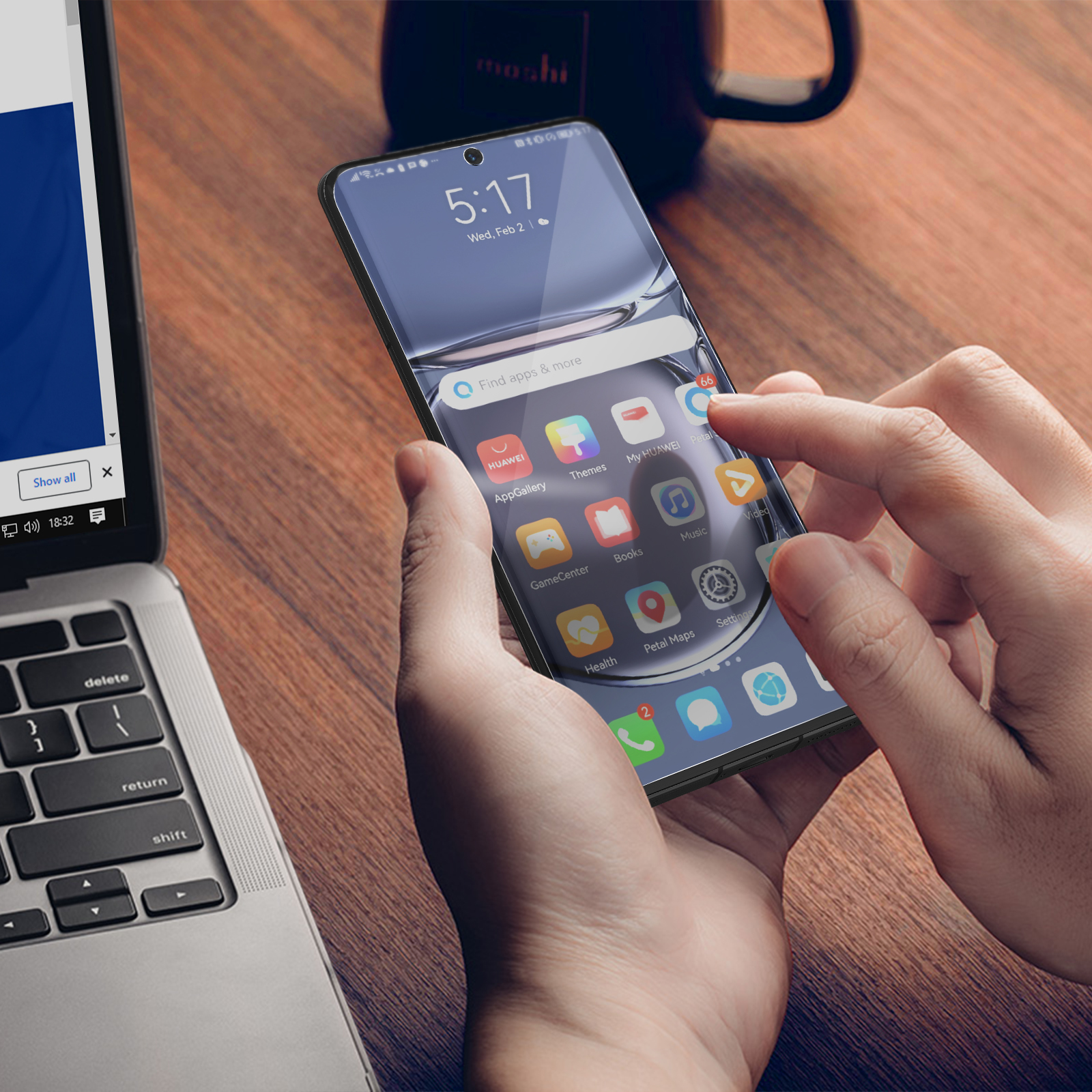 Ręka używa telefonu leżącego na drewnianym stole. Na stole są również laptop i kubek.