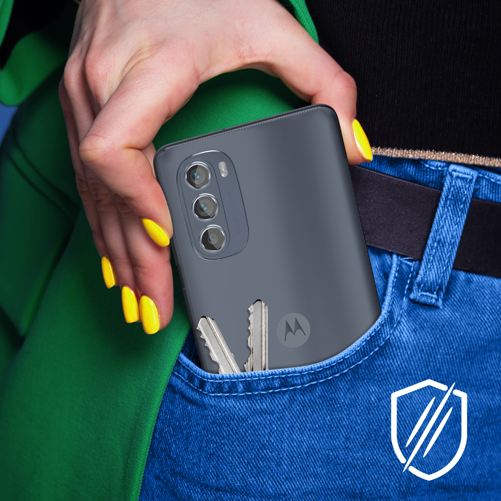 Une main aux ongles jaunes tient un téléphone gris. Des clés sont dans la poche d'un jean bleu. Une veste verte et une chemise noire sont visibles.