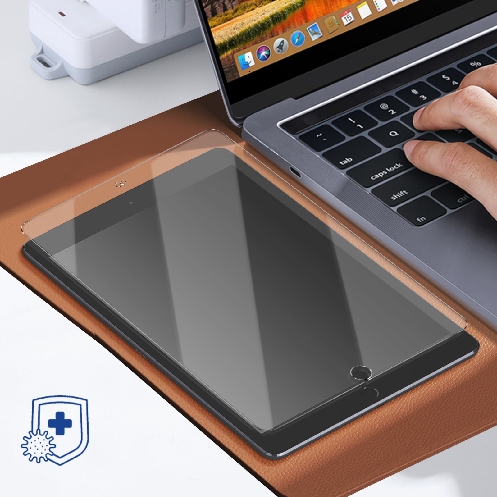 Tablet z ochraniaczem ekranu na biurku. Laptop w tle, ręka osoby na klawiaturze.