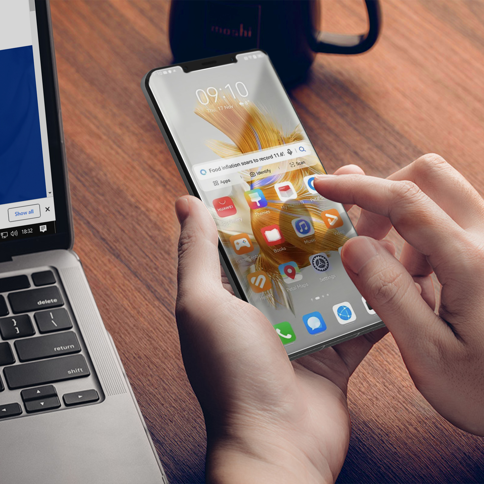 Una persona tiene in mano uno smartphone che mostra un risultato di ricerca di notizie. Un laptop è a sinistra, una tazza in alto a destra.
