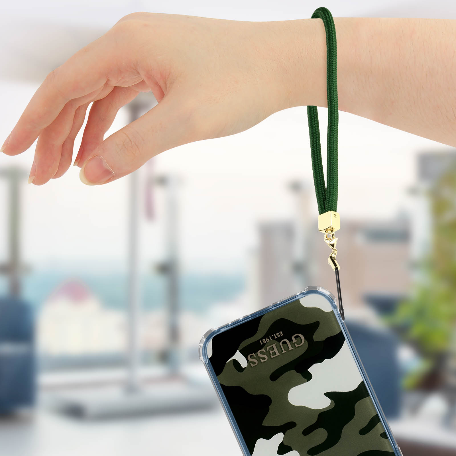 Mano che tiene il telefono con cinturino verde. Custodia mimetica visibile.