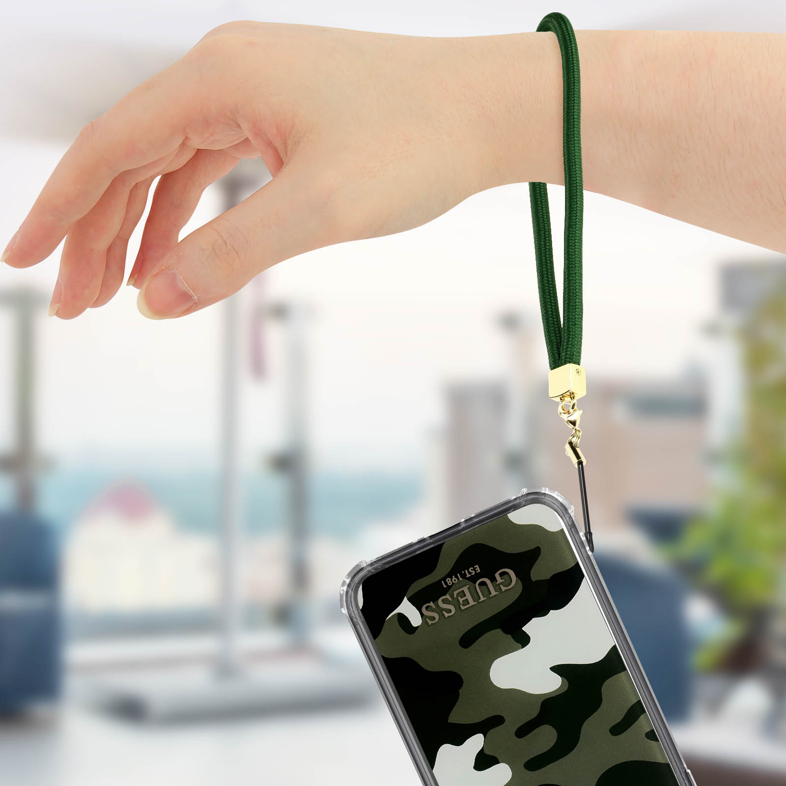 Hand mit grünem Handgelenkband, das ein Telefon mit Guess-Tarnmuster hält.
