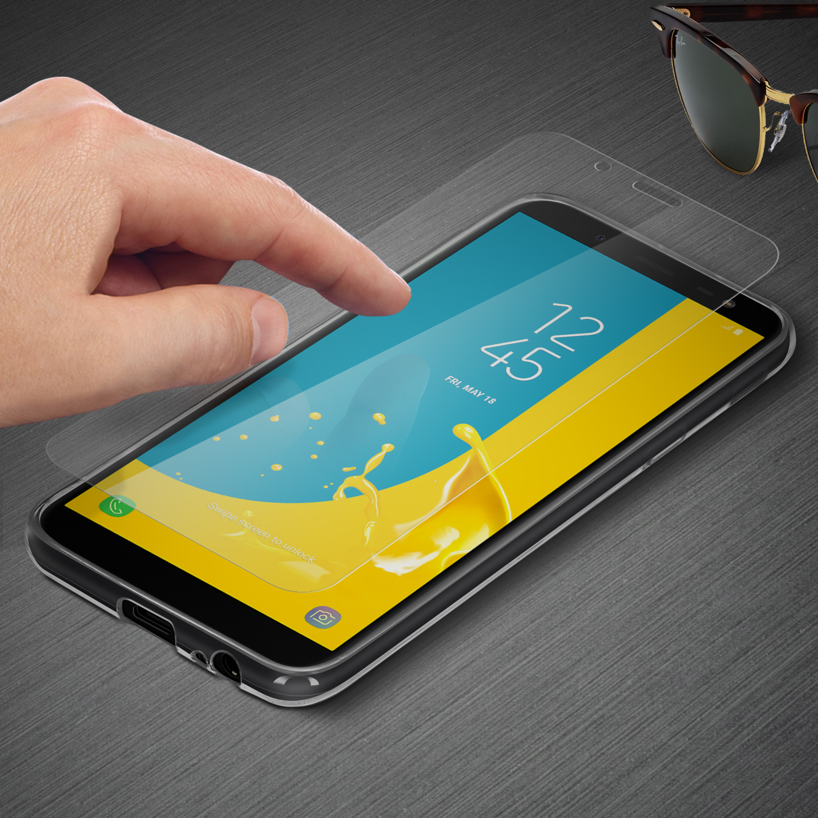Mano che applica una protezione schermo a un telefono, con occhiali da sole sullo sfondo, su una superficie grigia.