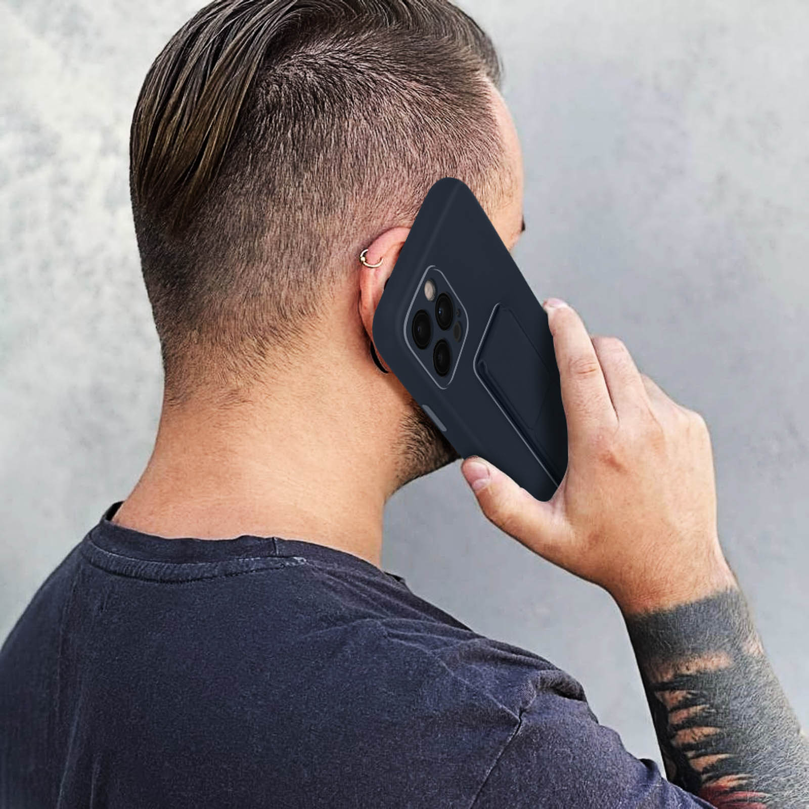 Mężczyzna trzymający telefon w granatowym etui. Ma kolczyk w uchu i ciemny t-shirt.