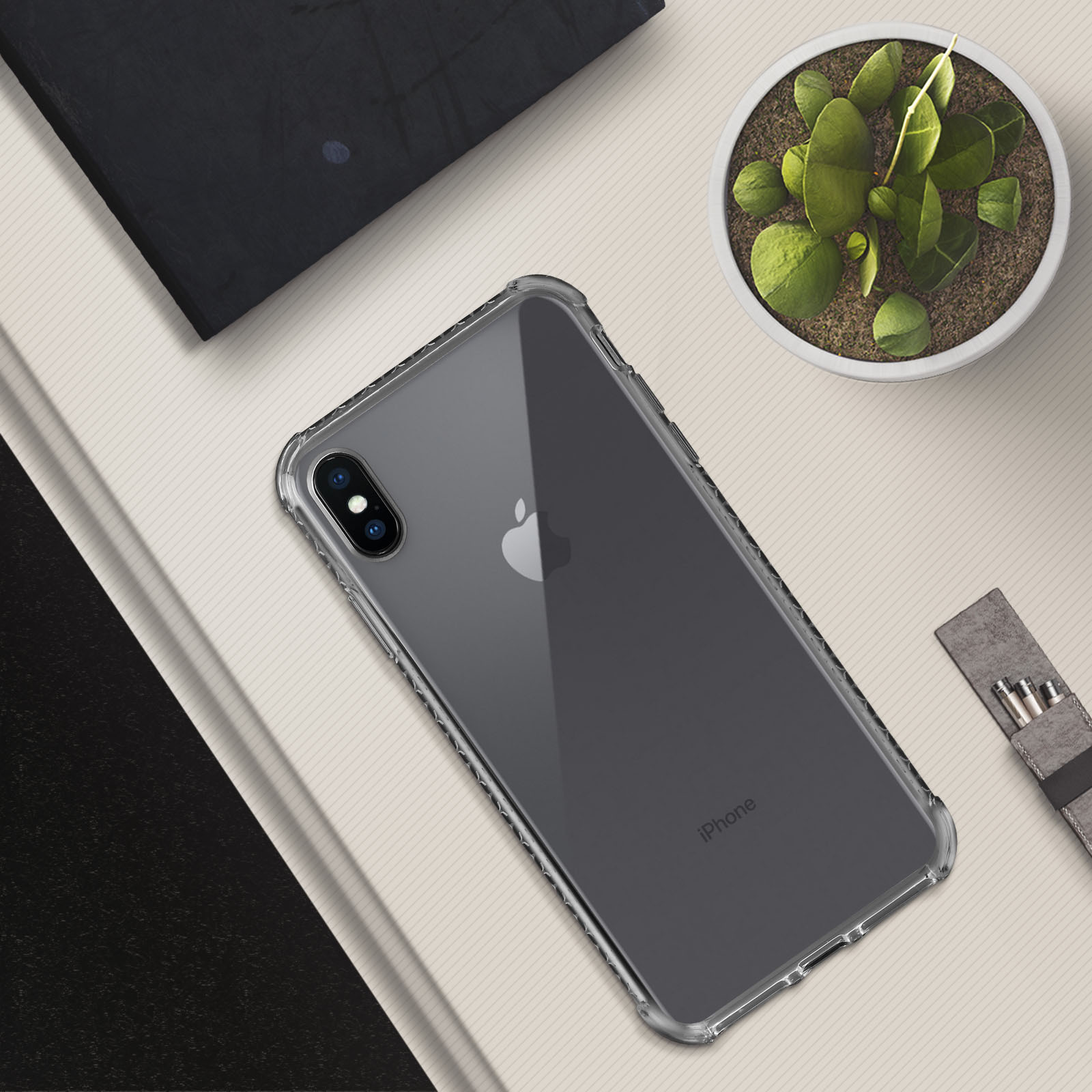Custodia trasparente per iPhone con telefono all'interno. Piccola pianta in alto a destra. Un taccuino nero in alto a sinistra.