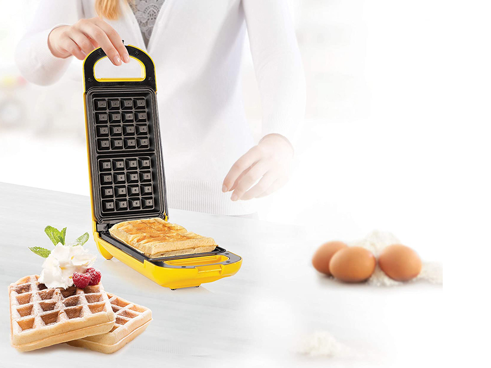 Wafflera gialla aperta, con waffle, uova e ingredienti su una superficie bianca.