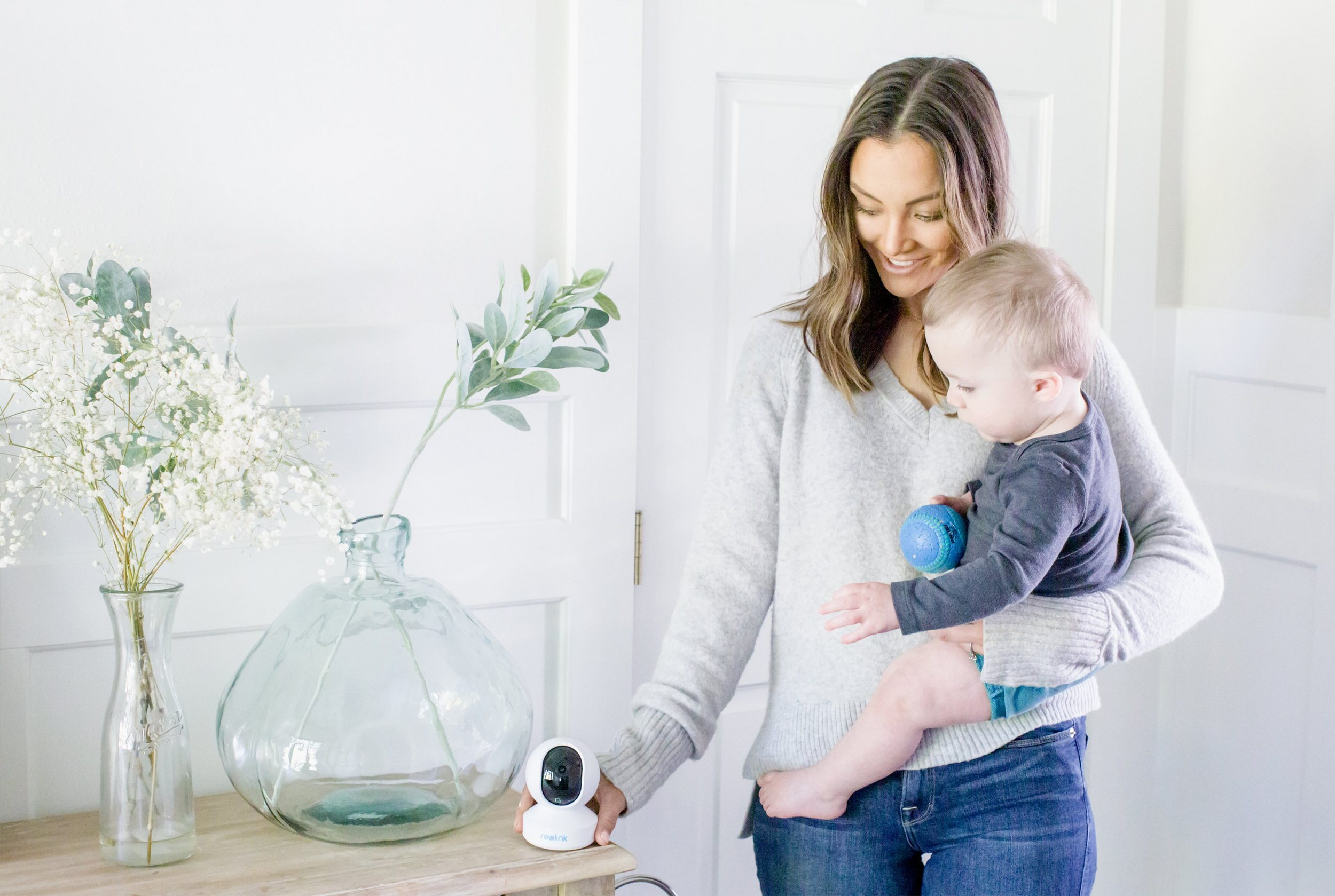 Frau hält ein Baby. Ein Babyphone steht auf einem Tisch. Blumen sind in einer Vase.