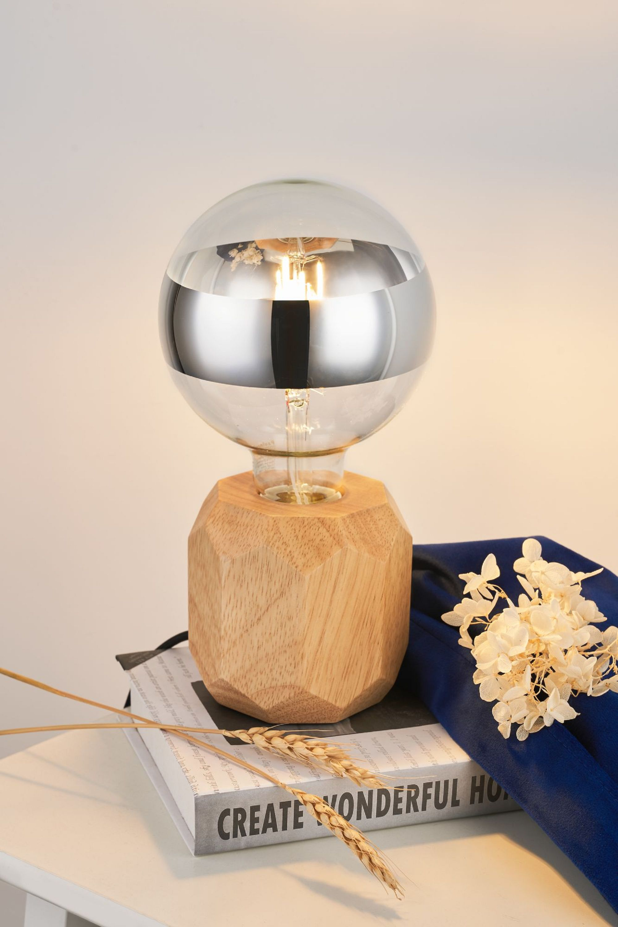 Eine Holzlampe mit einer Glühbirne. Die Glühbirne hat einen metallischen Look. Daneben liegen Bücher und Blumen.