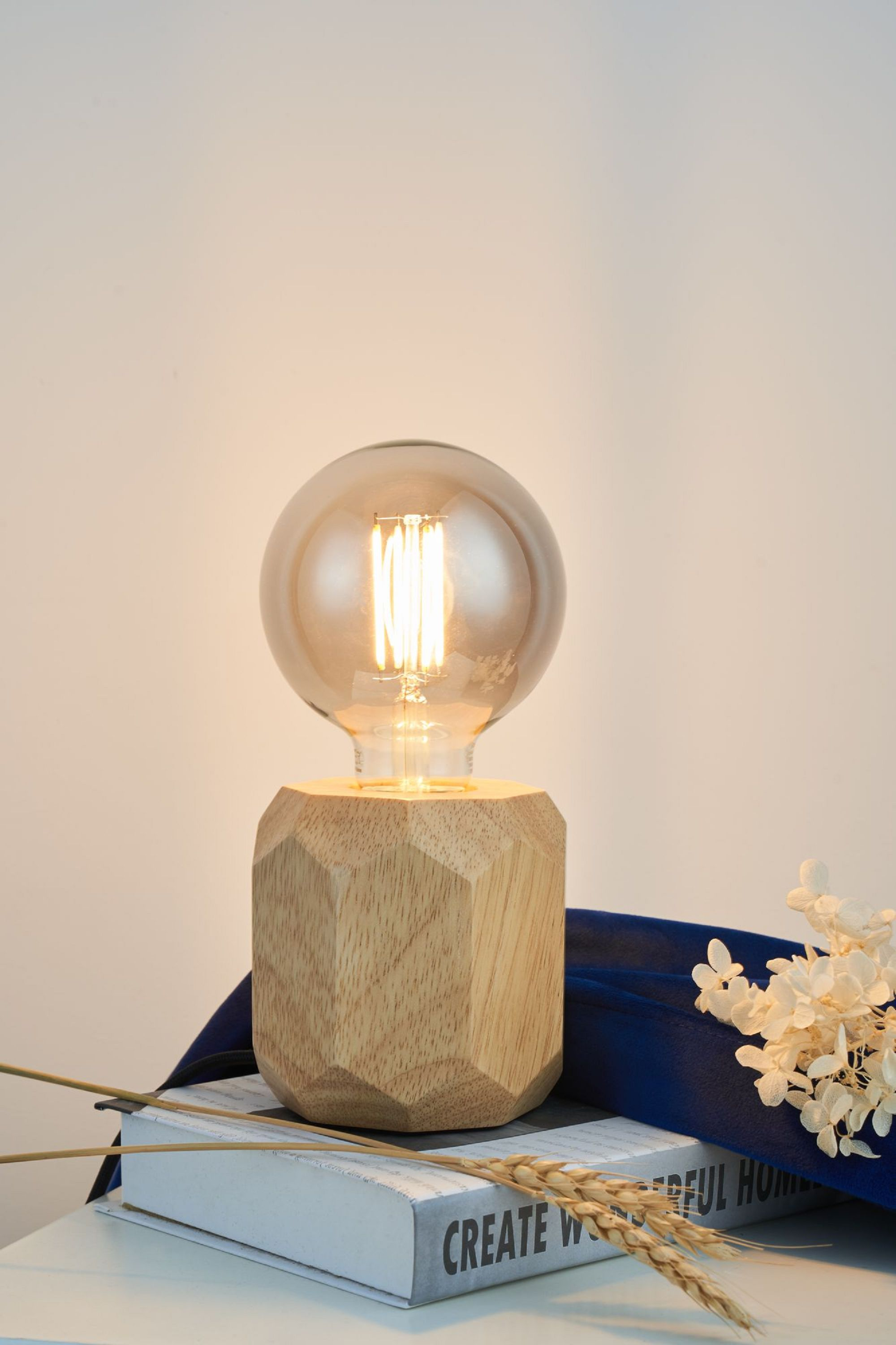 Eine Glühbirnenlampe steht auf einem Buch und blauem Tuch. Sie hat einen Holzfuß und eine Glühbirne.