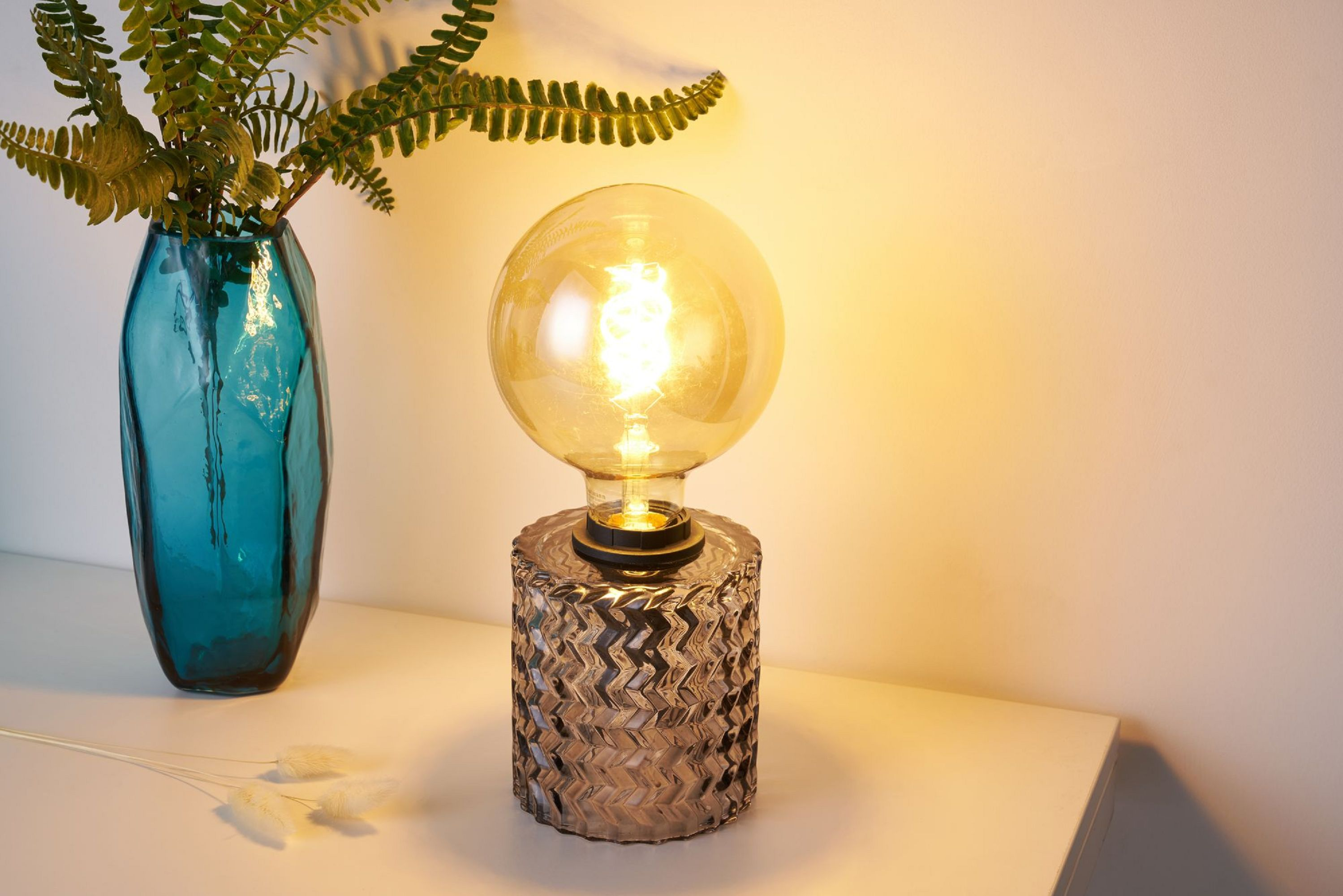 Lampa stołowa z okrągłą żarówką i wzorzystą podstawą. Paproć i wazon w tle, na białej powierzchni.