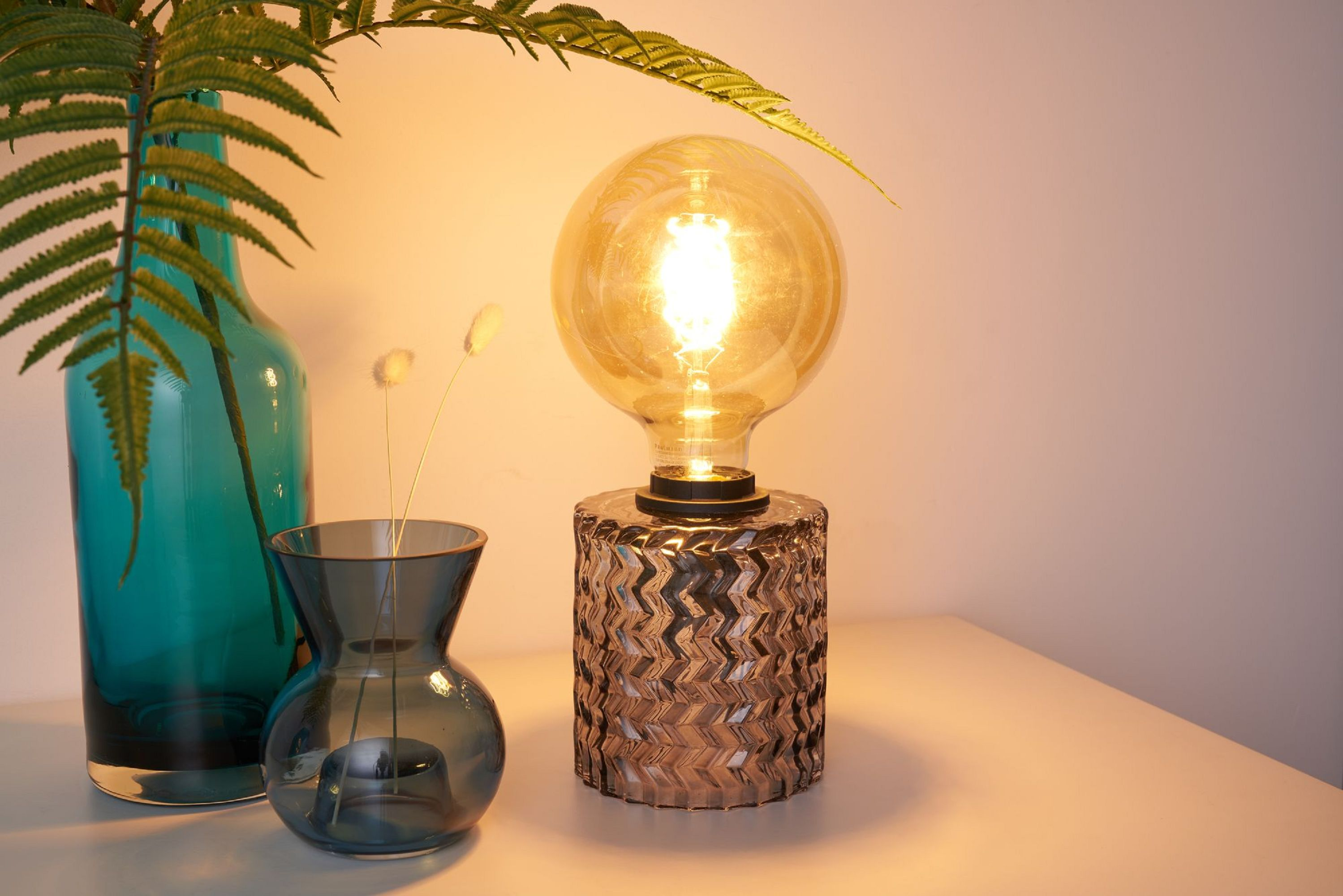 Lampa stołowa z dużą okrągłą żarówką. Wazon z paprocią i mniejszy wazon z suszonymi kwiatami.