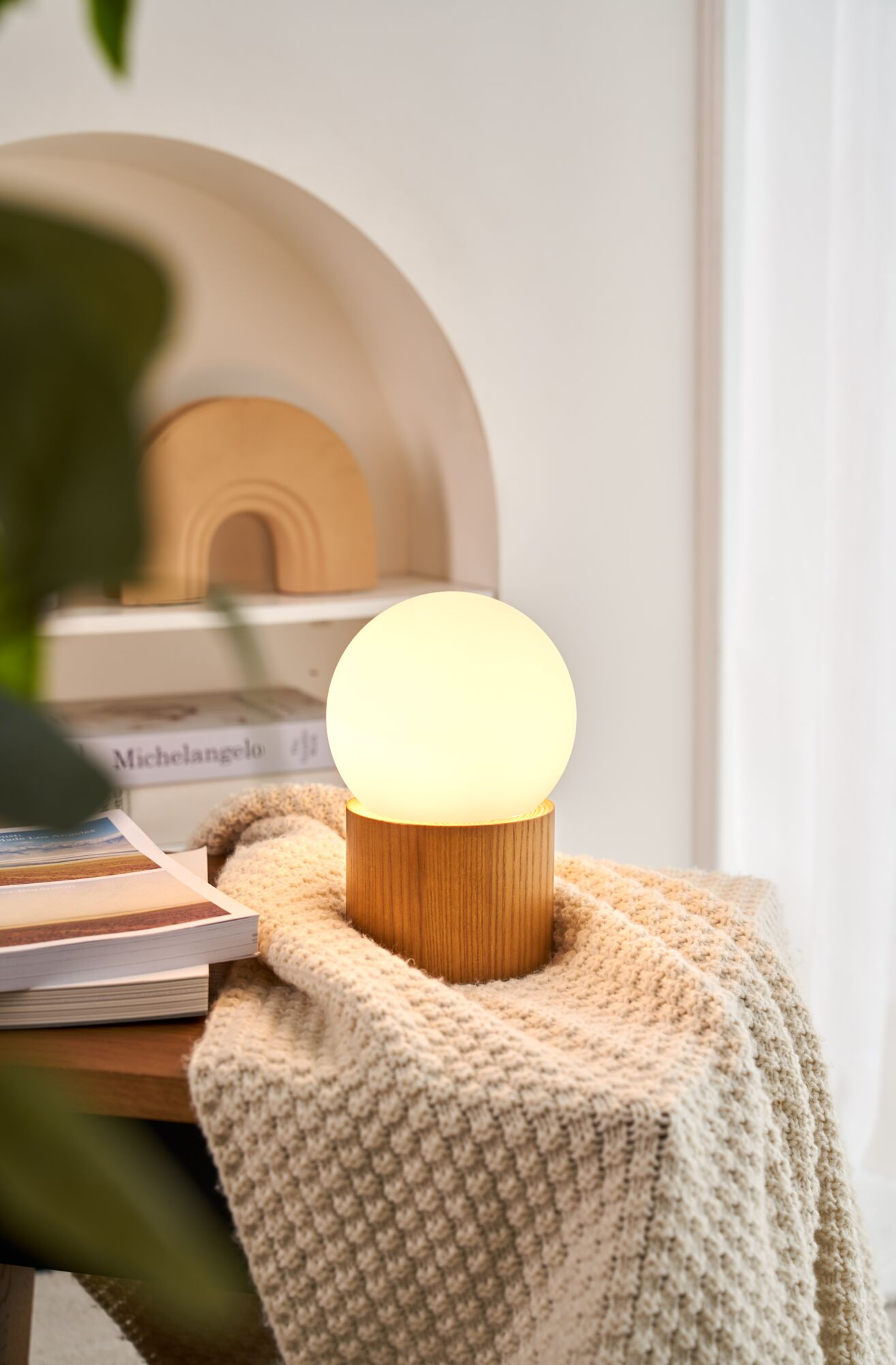 Oświetlona, okrągła, biała lampa stoi na drewnianej podstawie, na beżowym kocu, z książkami.