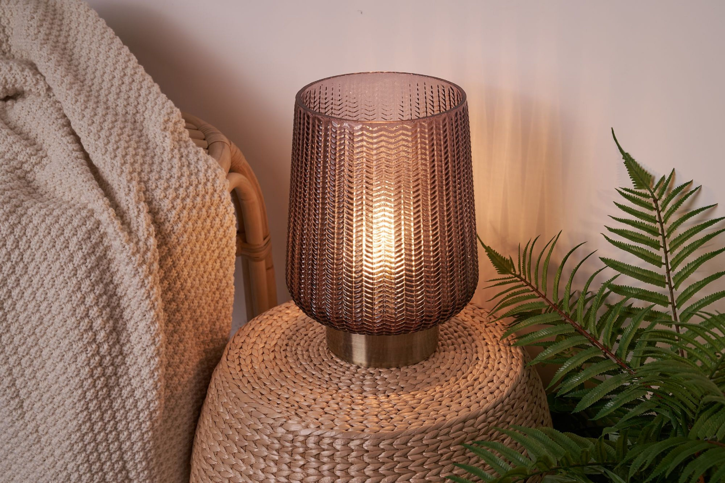 Eine Tischlampe mit pinkfarbenem Glasschirm und Messingfuß. Sie steht auf einem gewebten Tisch.
