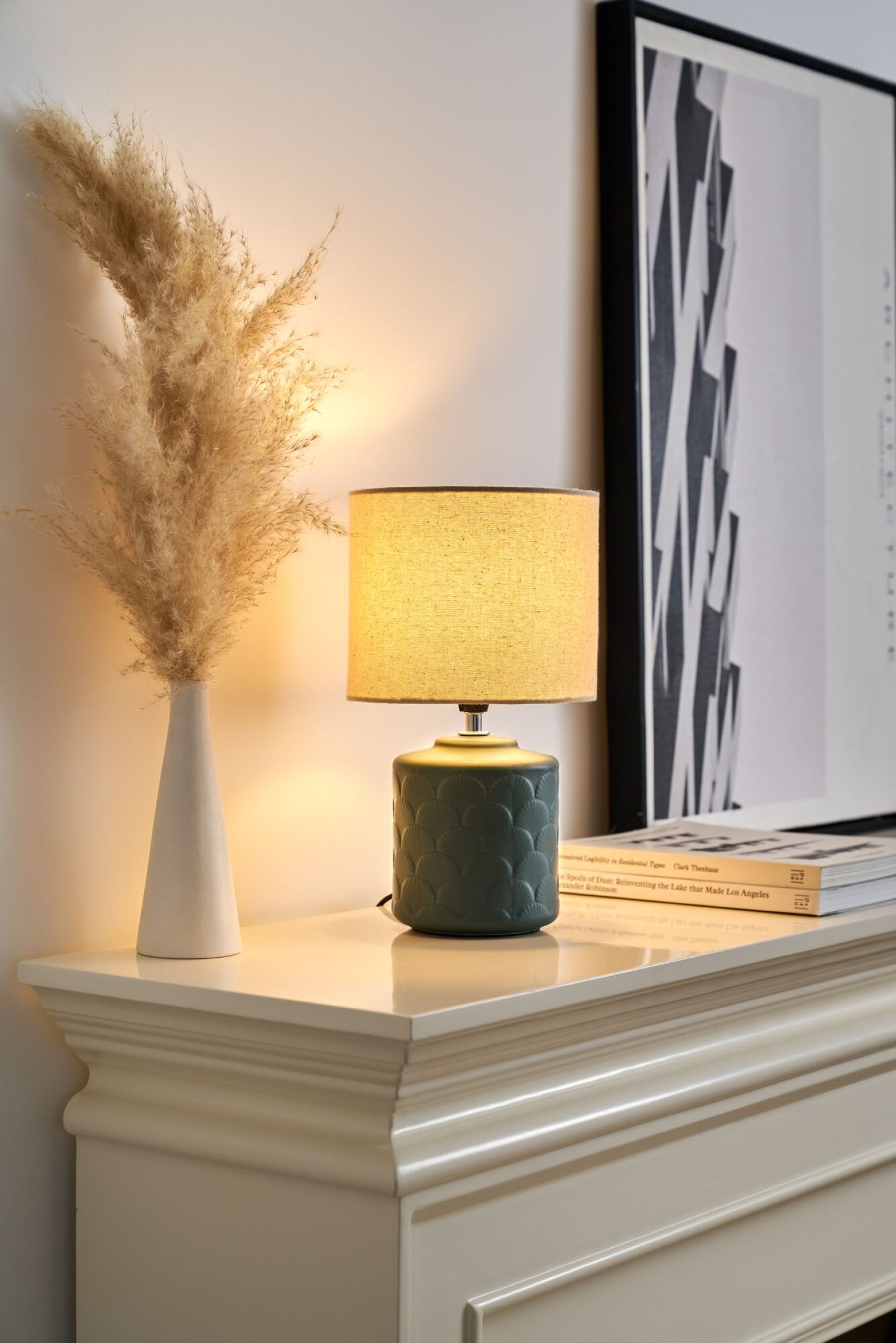Lampa na stole obok wazonu z trawą pampasową i oprawionym zdjęciem.