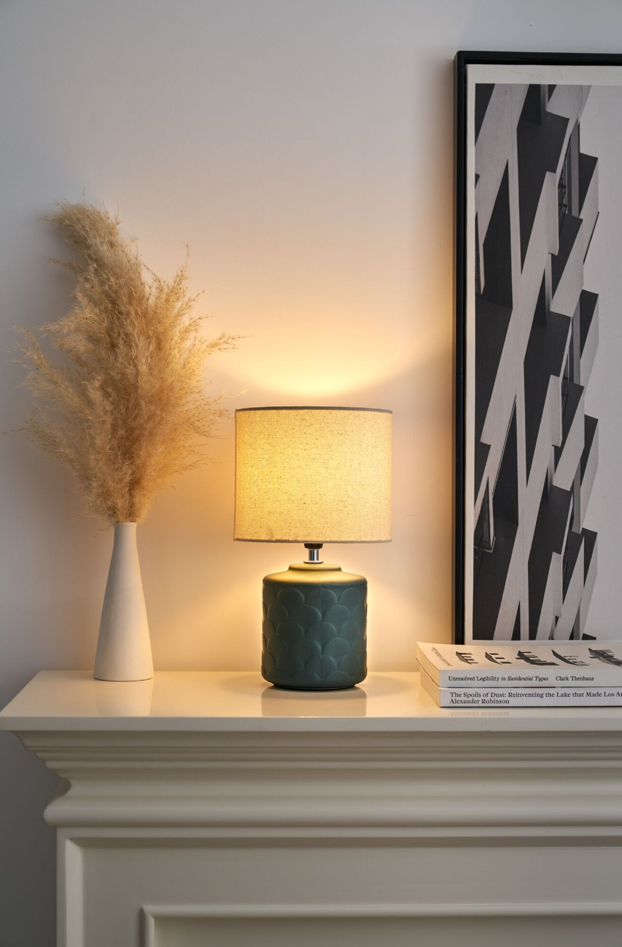 Lampa na białym kominku. Beżowy abażur, zielona podstawa, wazon z suszonymi kwiatami i książkami.