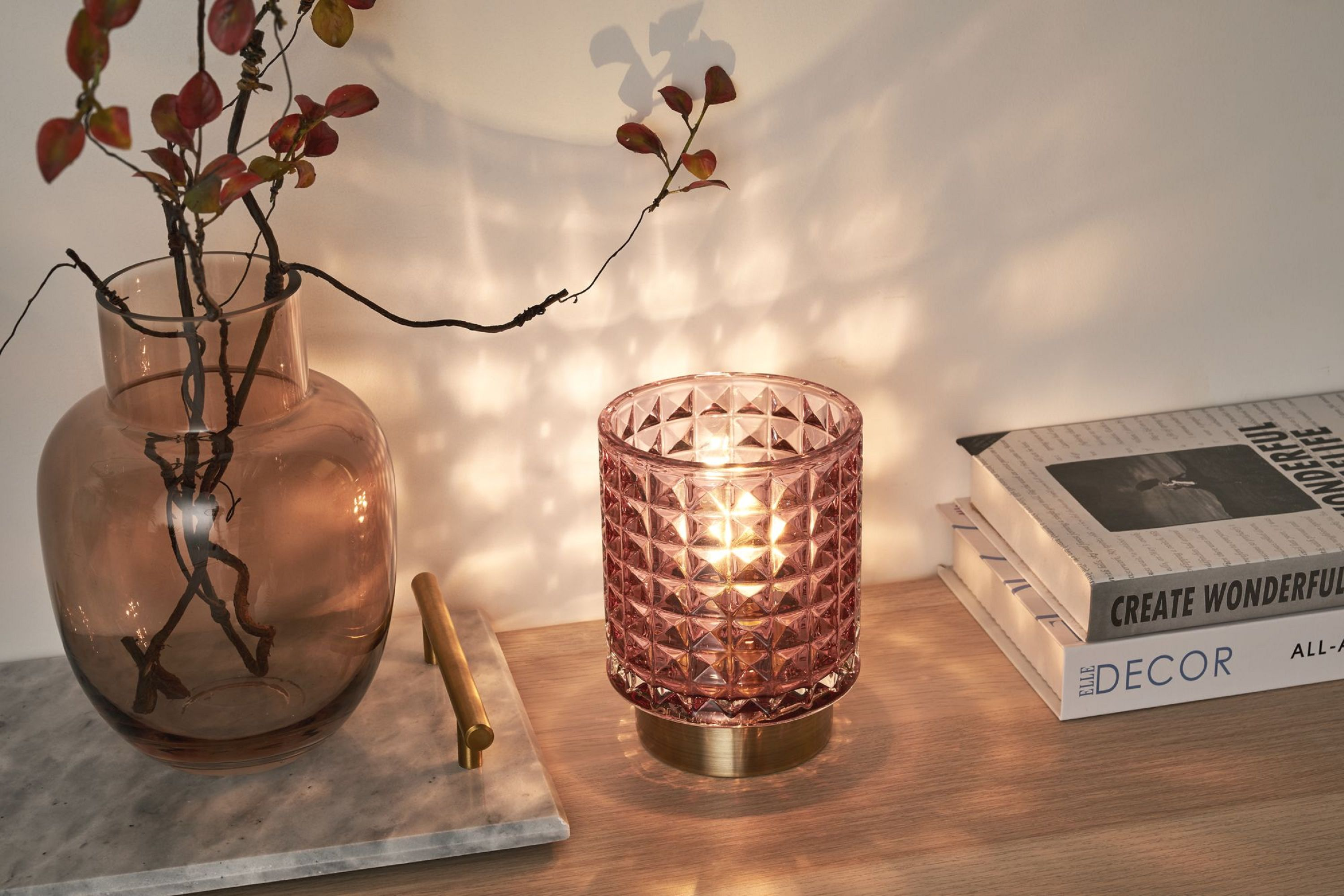 Różowa szklana lampa z zapaloną świecą, obok wazonu z czerwonymi gałęziami, na drewnie.