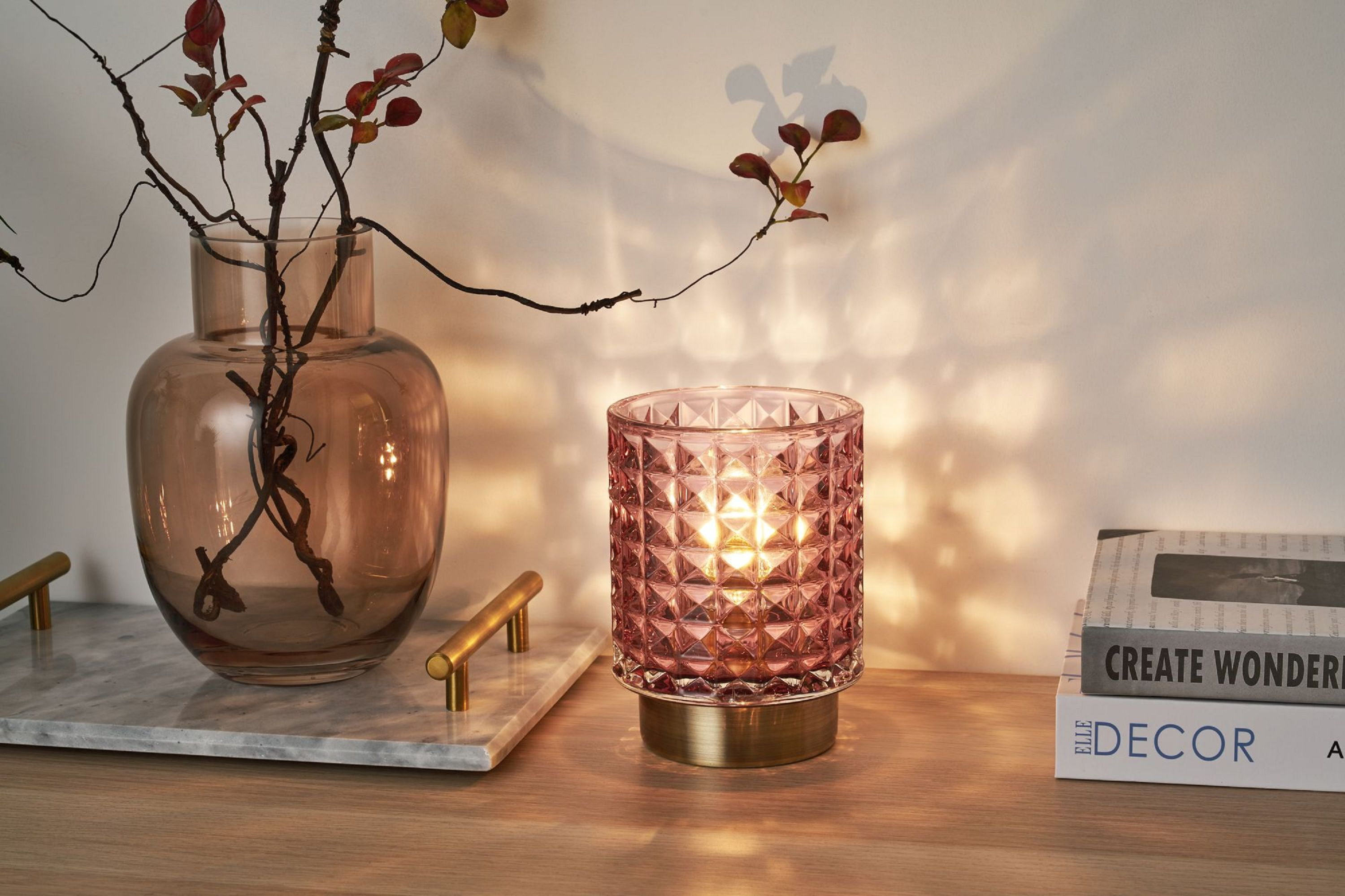 Różowy szklany świecznik na drewnianym stole. Wazon z gałęziami i książki w tle. Ciepłe oświetlenie.