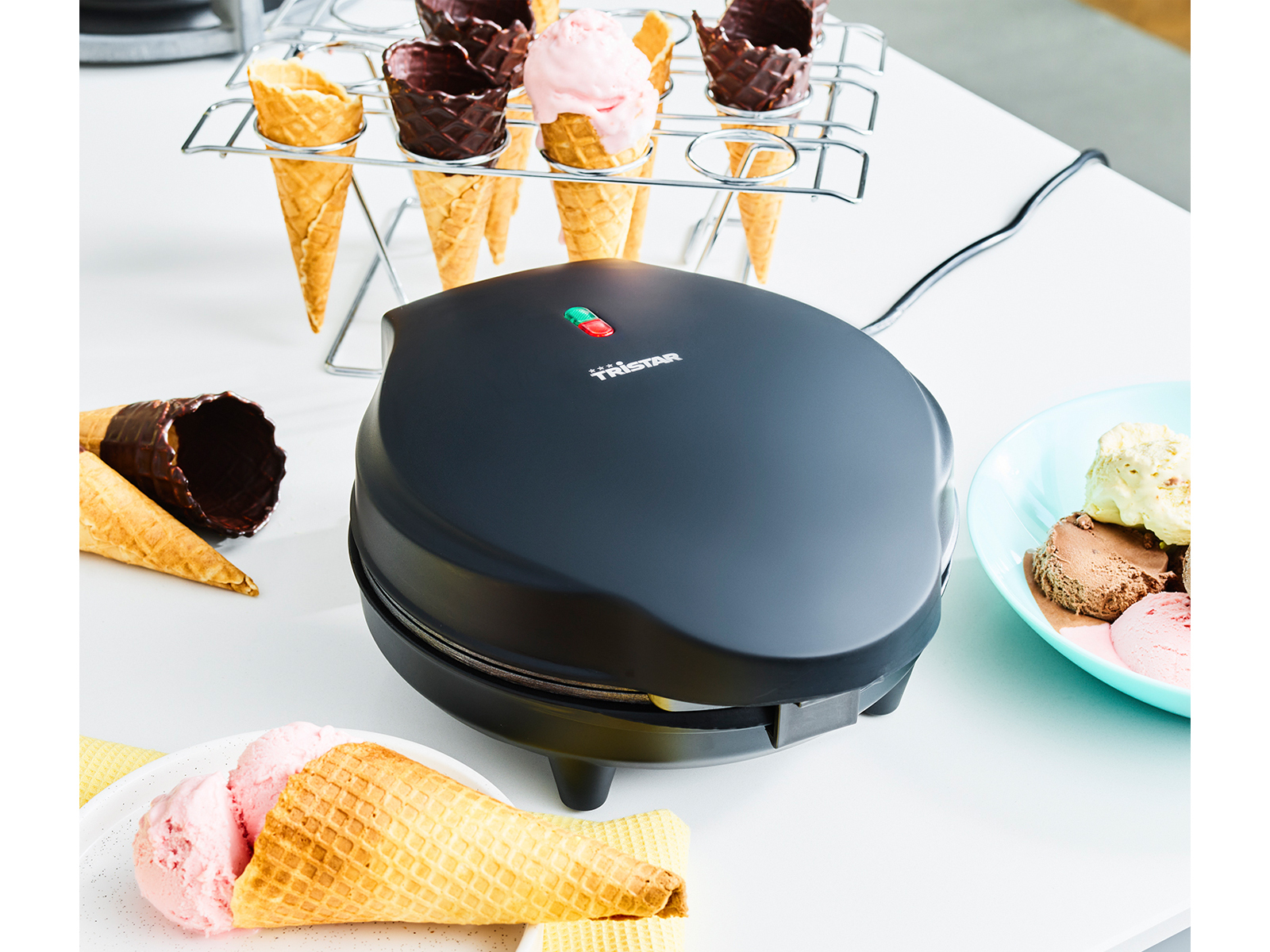 Schwarzer Waffeleisen mit Waffelhörnchen und Eisportionen. Weißer Tisch, sonniger Hintergrund.
