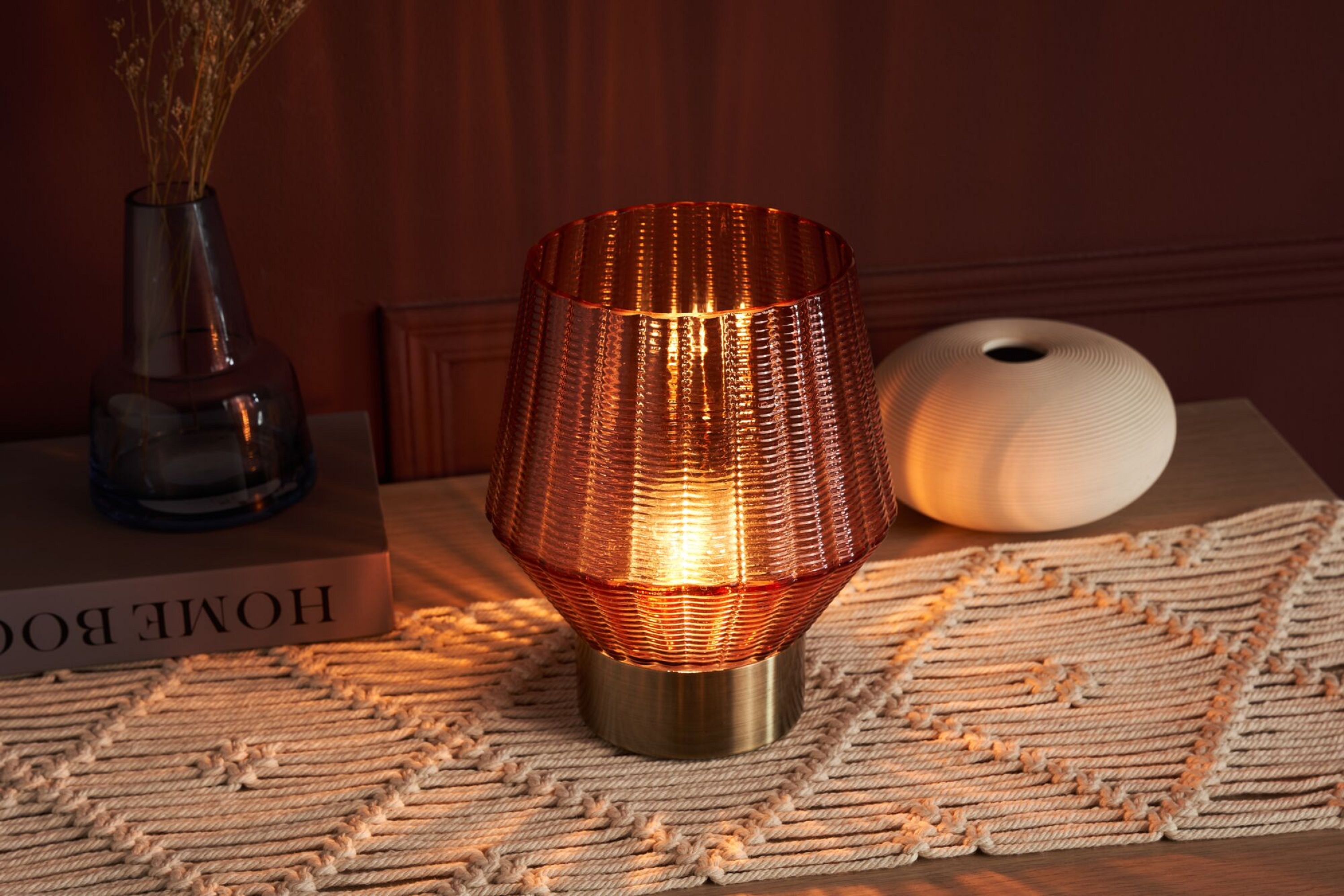 Rote Glaslampe mit Licht an, und eine weiße Vase, und ein Buch auf einem Holztisch.