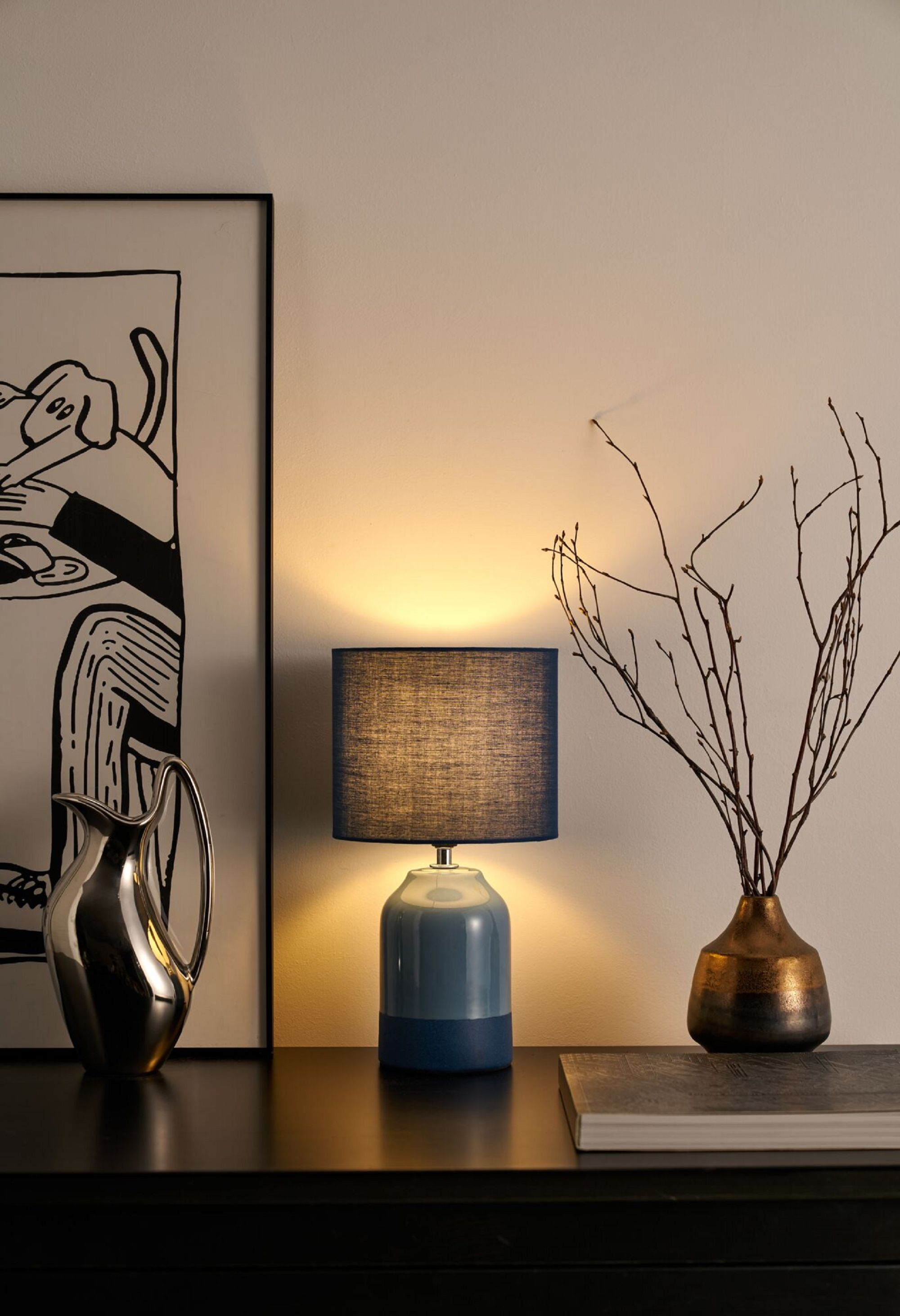 Niebiesko-szara lampa na czarnym stole, obok srebrnego dzbanka i wazonu z gałęziami.