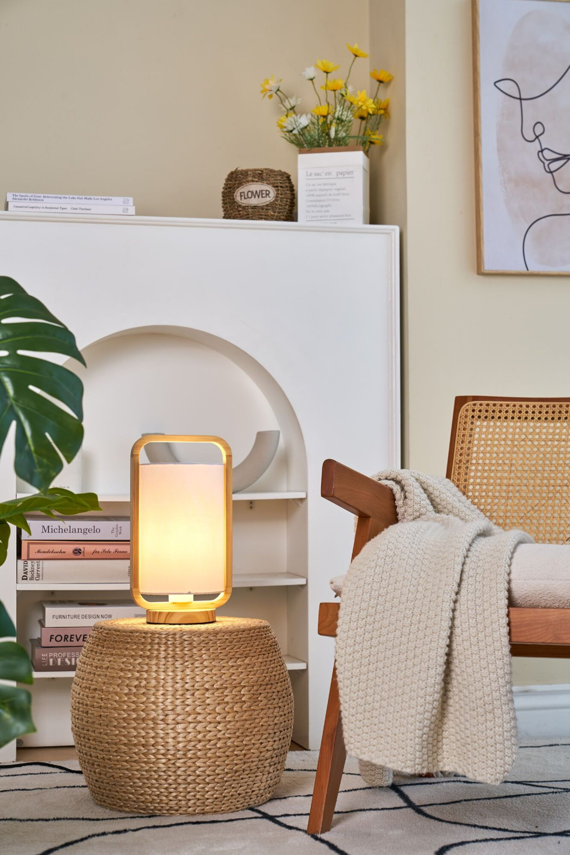 Drewniana lampa na wiklinowym stoliku. Krzesło z kocem po prawej. Książki i dekoracje w tle.