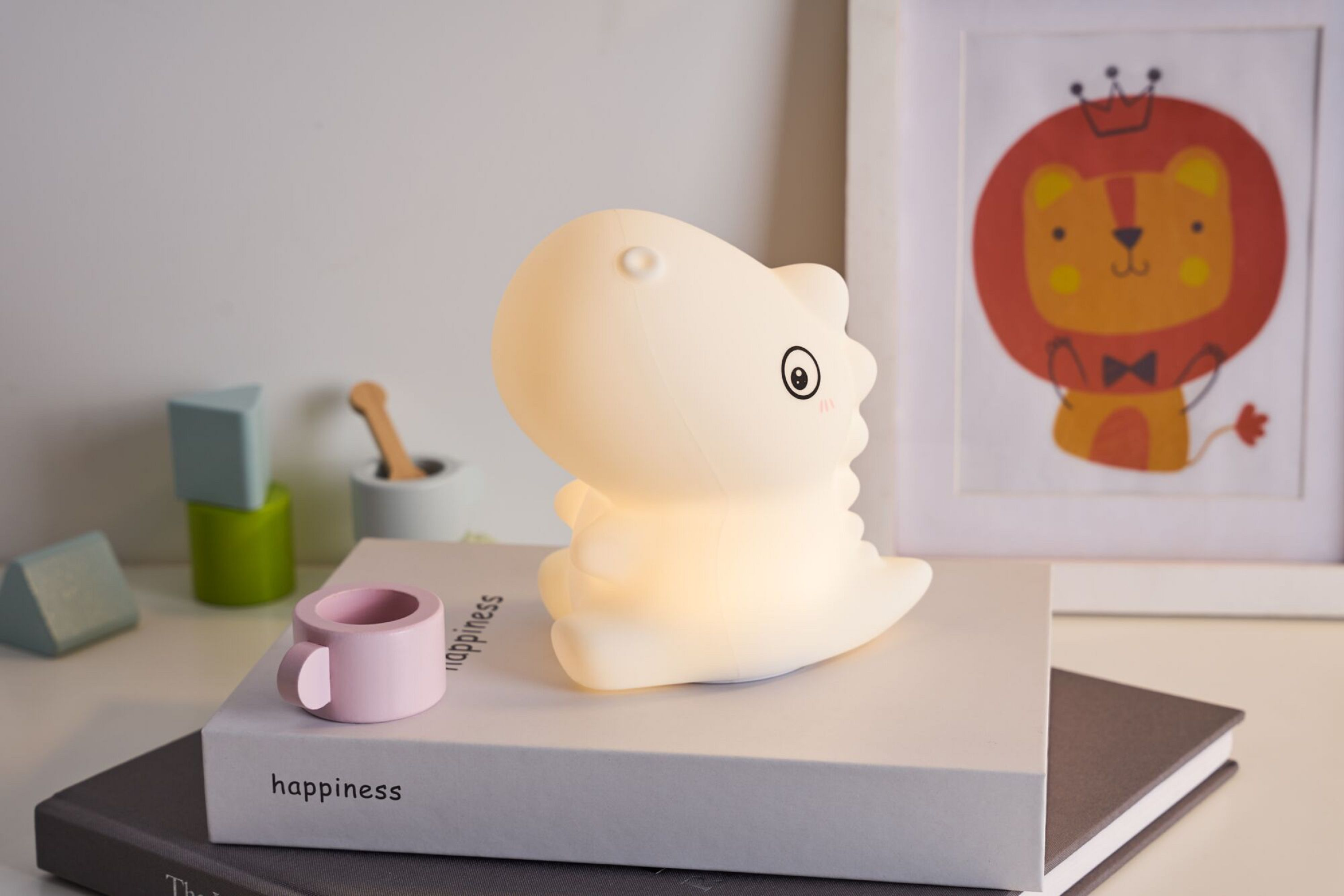 Weiße Dinosaurierlampe auf einem Bücherstapel, mit einer rosa Tasse und einem Löwenbild.