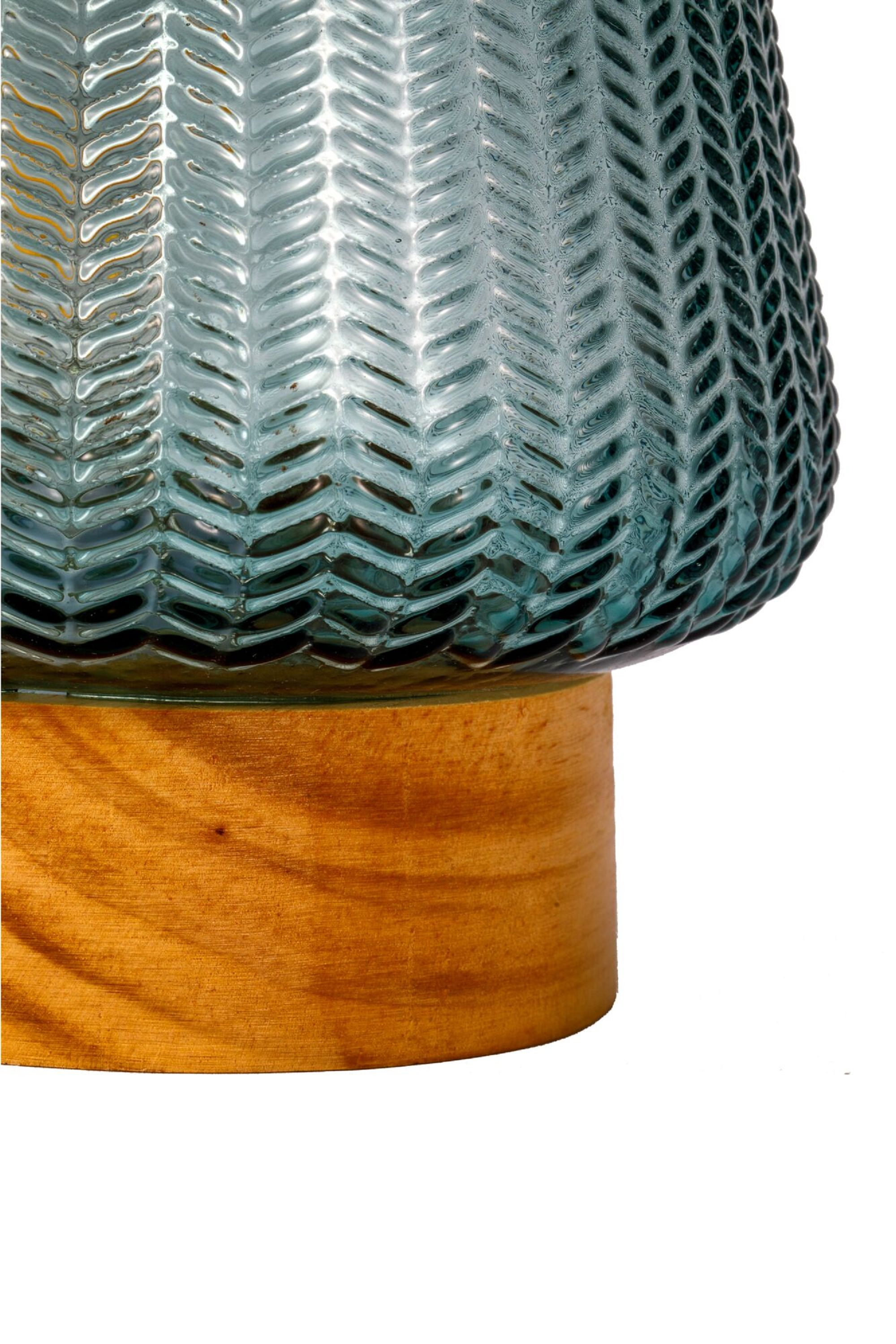 Niebieska szklana lampa z drewnianą podstawą. Szkło ma wzór jodełki. Białe tło.