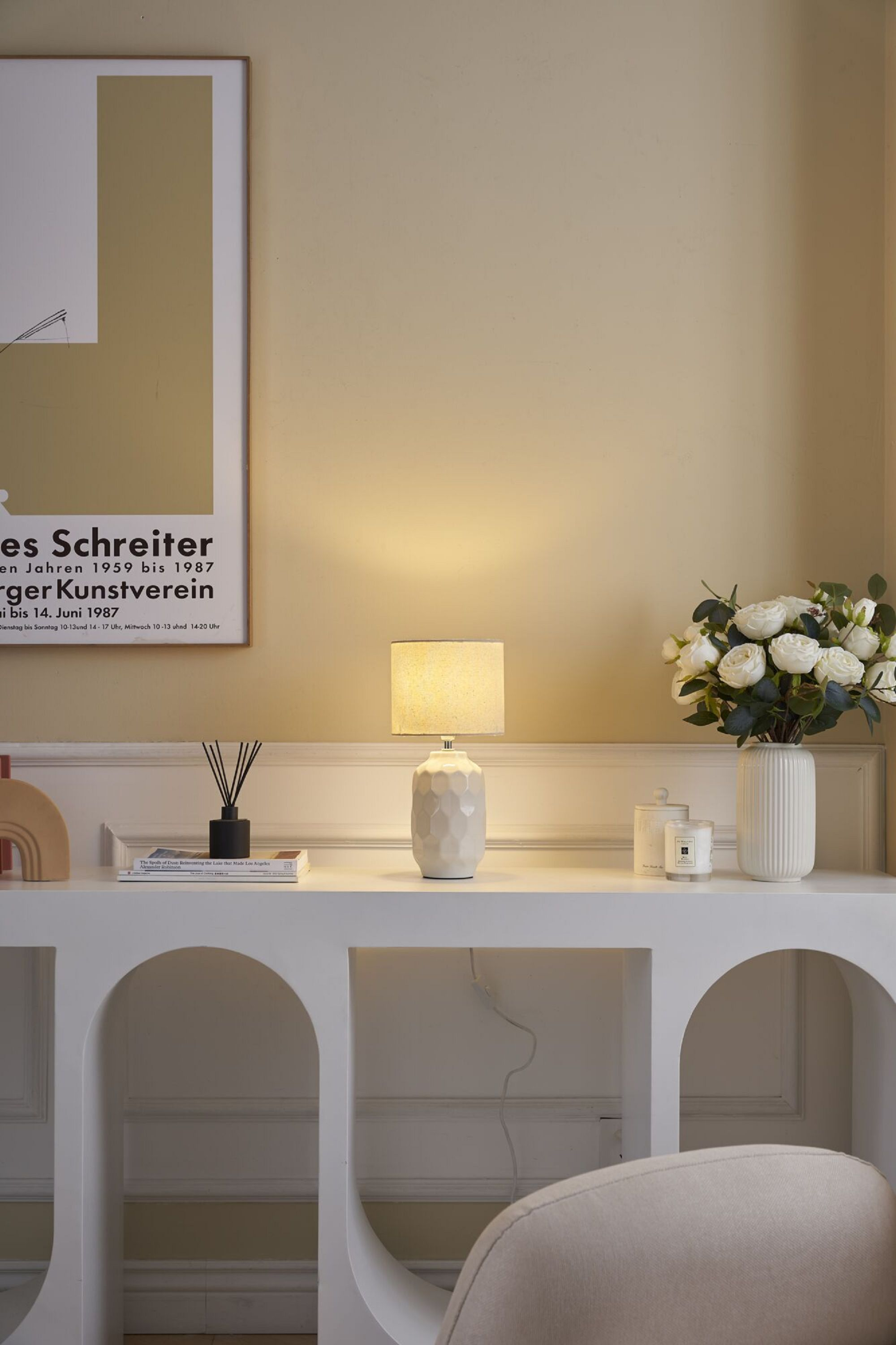 Lampa na białej konsoli z kwiatami i świecami, grafika na ścianie i krzesło w tle.