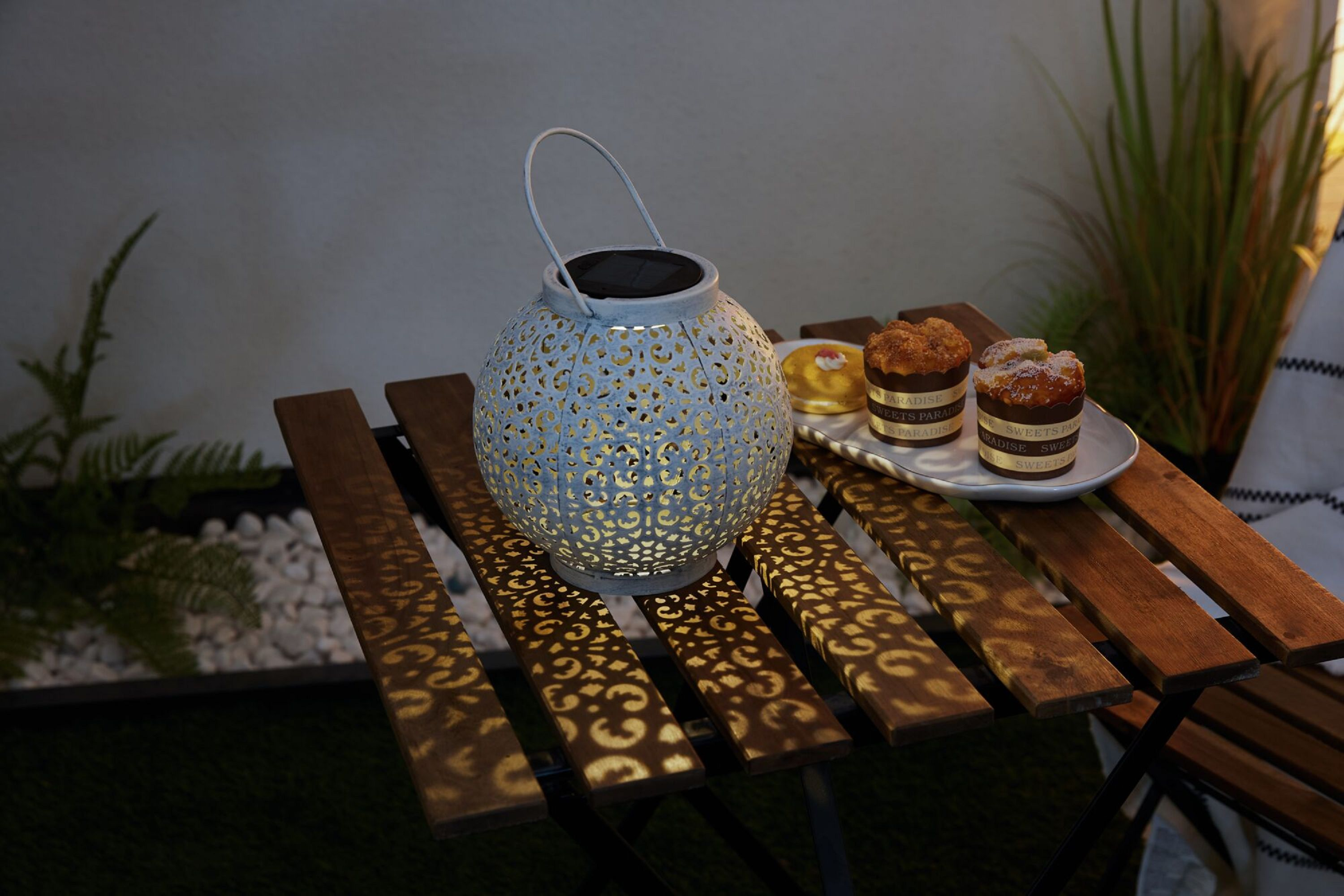 Eine weiße Laterne auf einem Holztisch mit kleinen Kuchen, beleuchtet vom Laternenlicht.