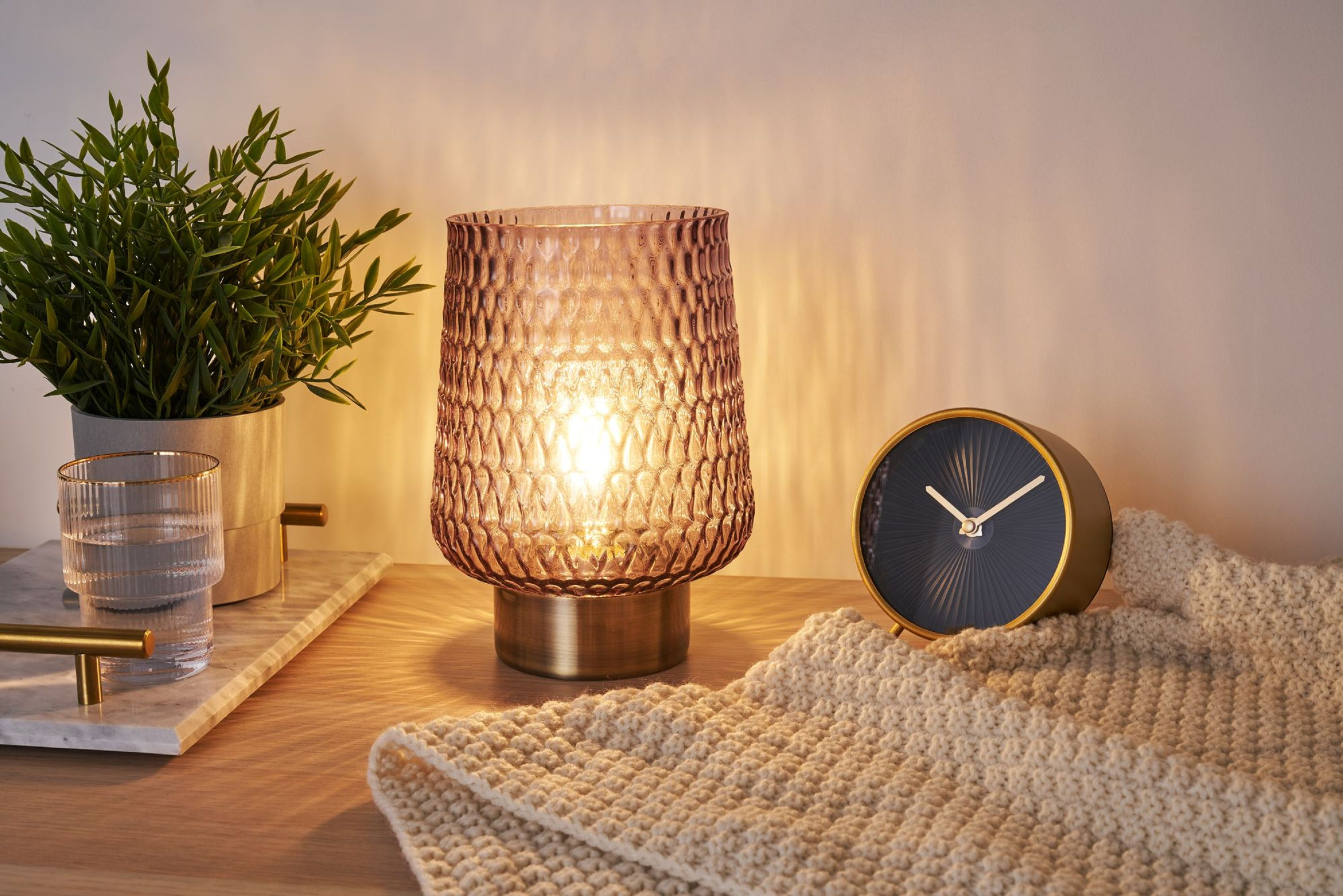 Różowa szklana lampa o teksturowanej powierzchni, książka, zegar i roślina na drewnianym stole.